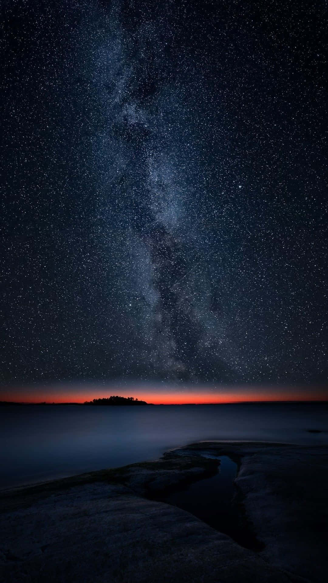 手機 桌布 壁紙 — 一個令人嘆為觀止的夜空，佈滿星星和銀河，拱形在平靜的海洋和遠處的島嶼輪廓之上。