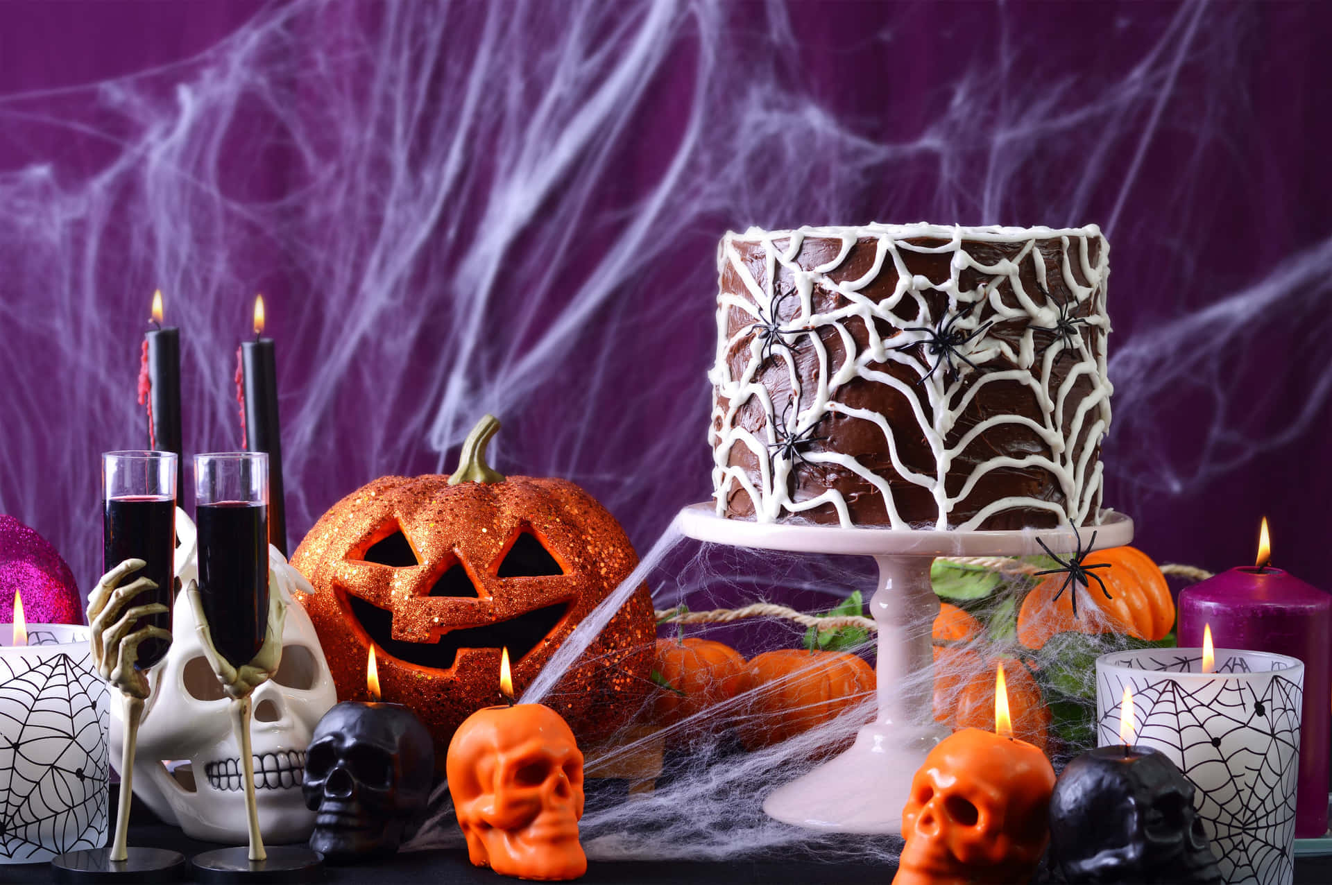 Halloween Props 桌布 壁紙 — 一個令人毛骨悚然的萬聖節甜品桌，上面有蜘蛛網蛋糕、發光南瓜和骷髏蠟燭，背景是紫色蜘蛛網。