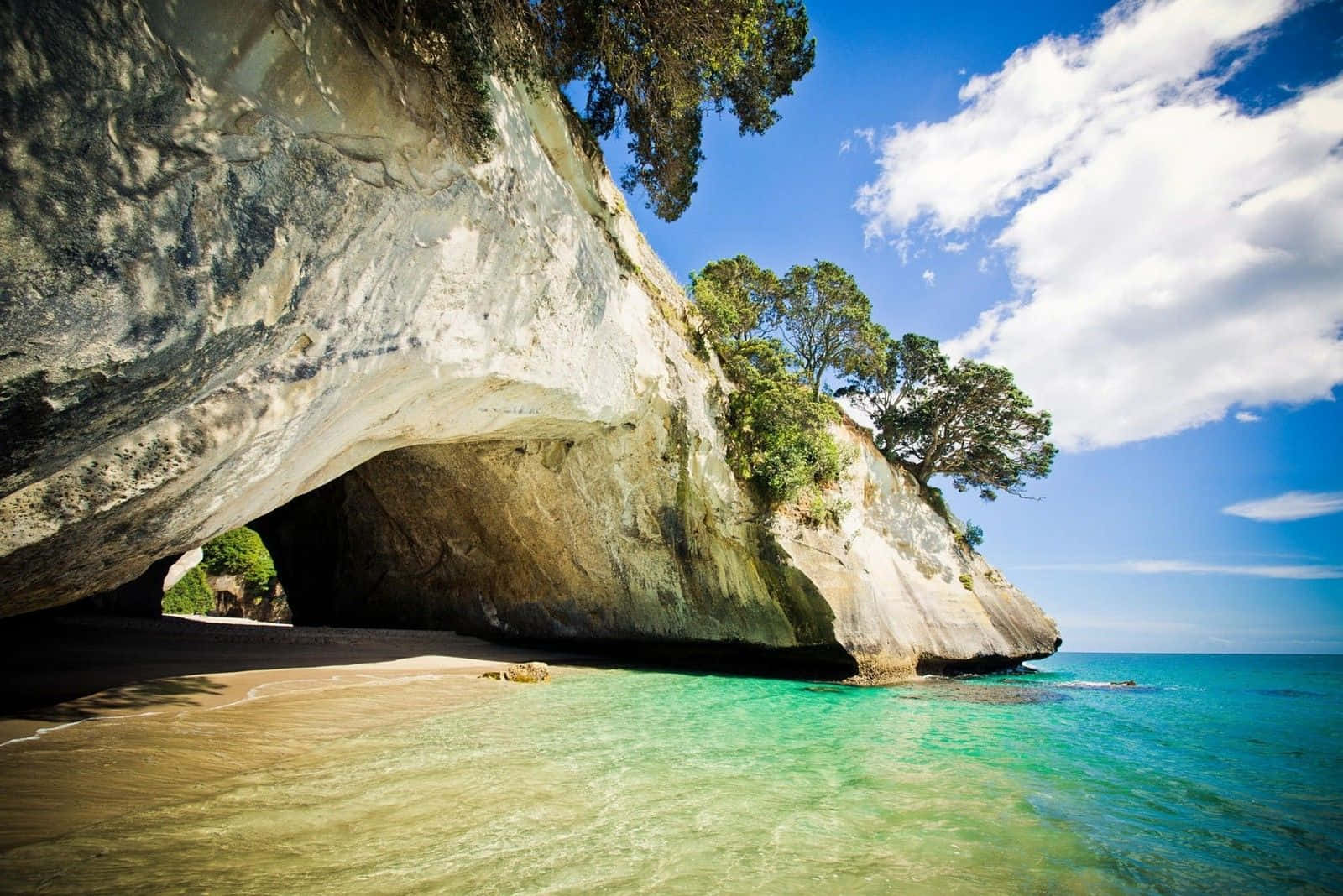 New Zealand 桌布 壁紙 — 一個令人驚嘆嘅天然海蝕洞，有藍綠色海水同沙灘，上面係晴朗藍天同 fluffy 雲。