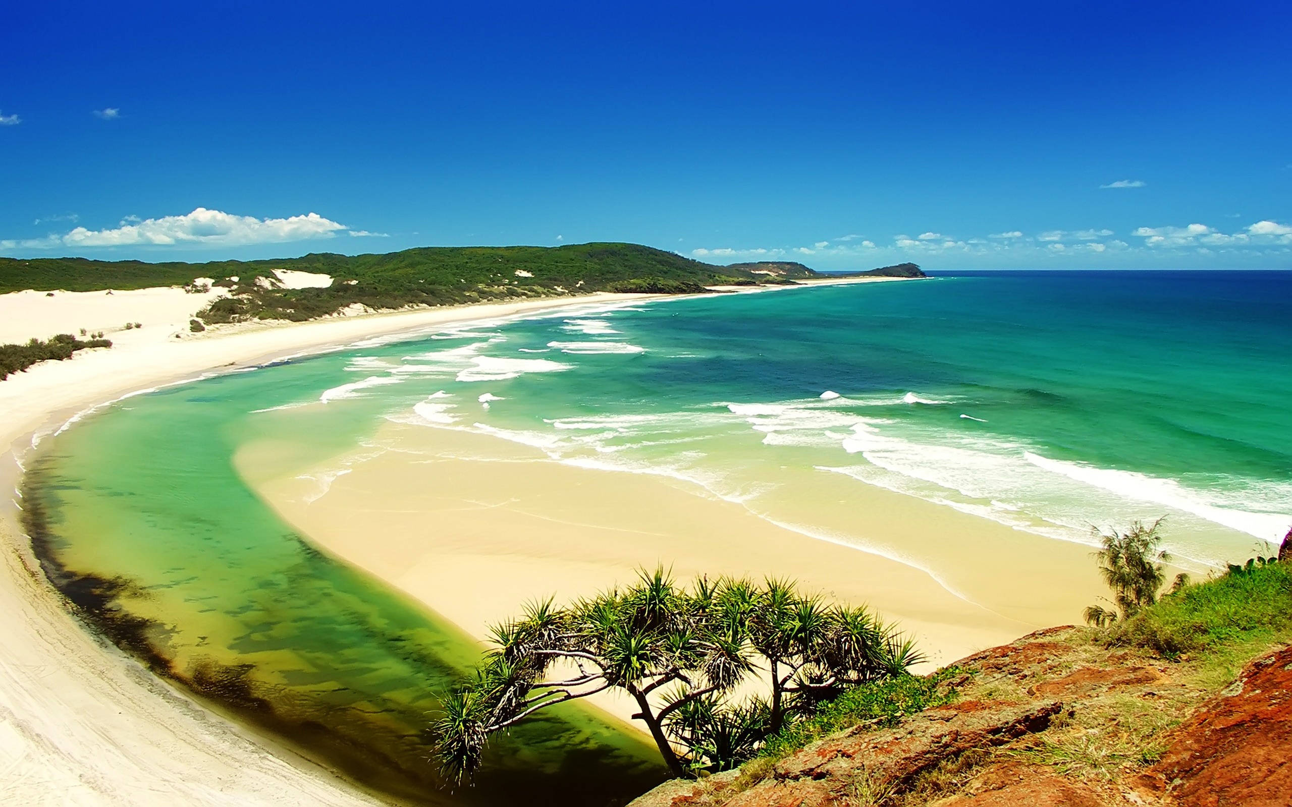 Beach Desktop 桌布 壁紙 — 一個令人驚嘆的彎曲海灘，擁有碧綠的海水和白色的沙灘，在明亮的藍天下被鬱鬱蔥蔥的綠色山丘環繞。