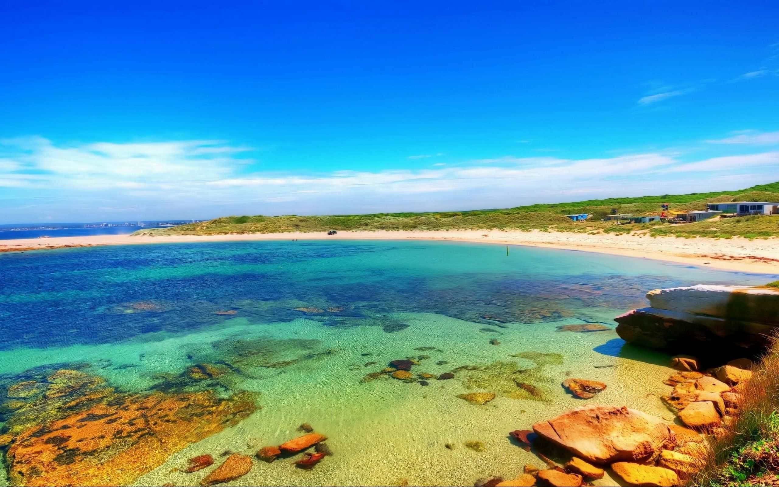 Australia Beach 桌布 壁紙 — 一個令人驚嘆的海岸小海灣，清澈的藍綠色海水顯露出色彩繽紛的岩石和沙灘，上方是明亮的藍天。
