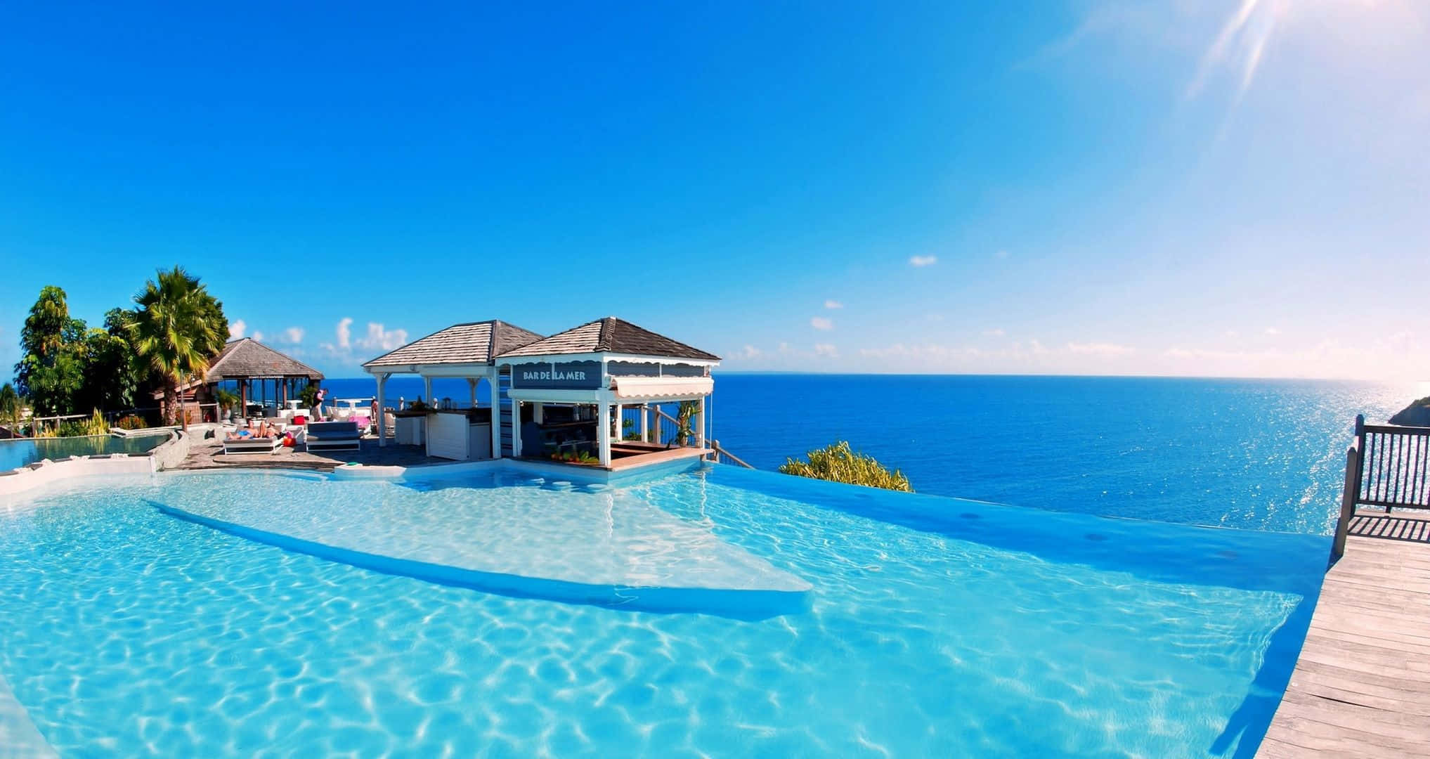 Beach Resort 桌布 壁紙 — 一個令人驚嘆的無邊際泳池俯瞰著廣闊的藍色海洋，在晴朗的天空下，附近有棕櫚樹和茅草屋頂酒吧。