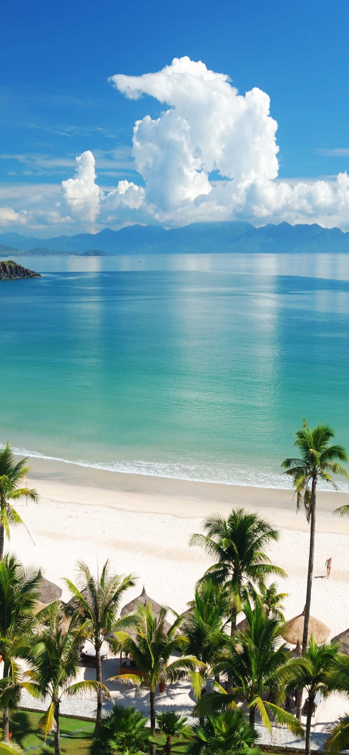 iPhone X Beach 桌布 壁紙 — 一個令人驚嘆的熱帶海灘，擁有碧綠的海水、白色的沙灘和棕櫚樹，在明亮的藍天下有蓬鬆的雲朵。
