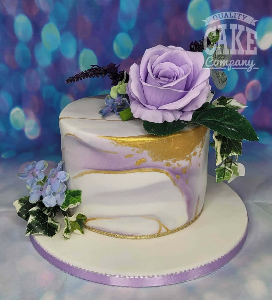 Floral Cake 桌布 壁紙 — 一個令人驚嘆的紫色和金色大理石蛋糕，上面點綴著一朵大薰衣草玫瑰和精緻的花朵，背景是充滿活力的散景。