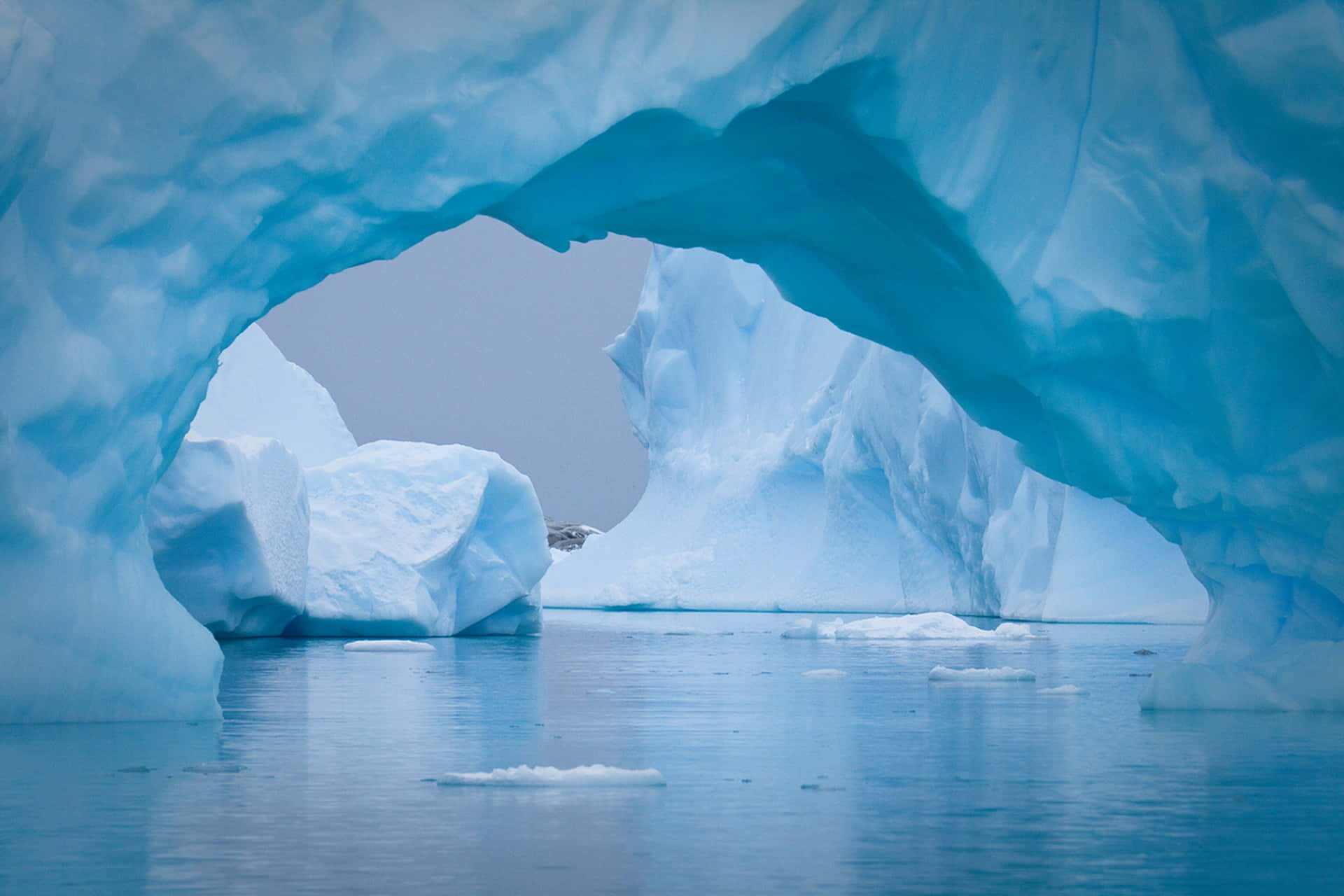 Antarctica 桌布 壁紙 — 一個令人驚嘆的藍色冰拱門框住了寧靜的冰川景觀，平靜的水面和漂浮的冰山。