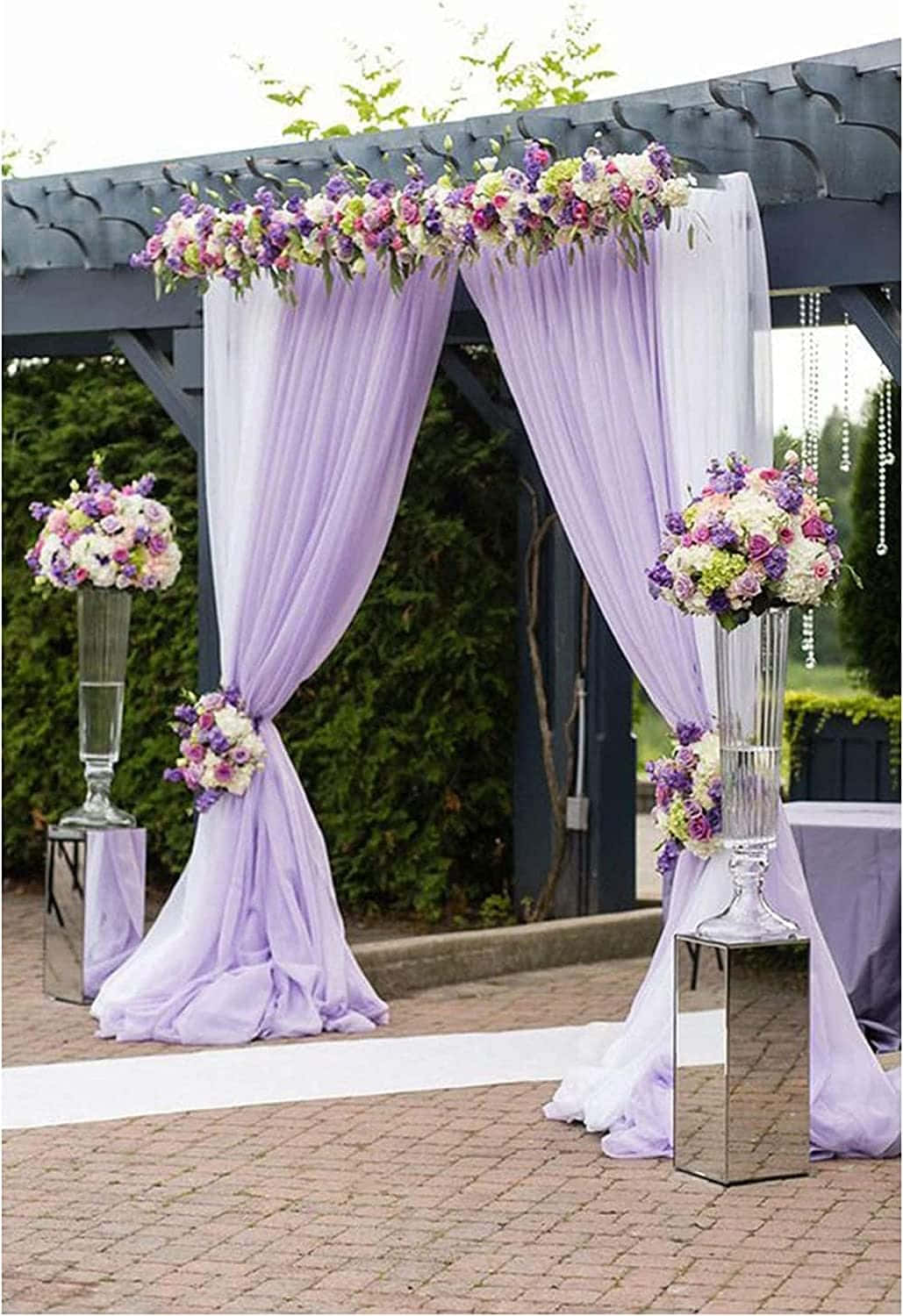 Garden Wedding 桌布 壁紙 — 一個以薰衣草覆蓋的婚禮拱門，裝飾著紫色、白色和綠色的花卉佈置，矗立在磚砌庭院中，周圍環繞著綠植。
