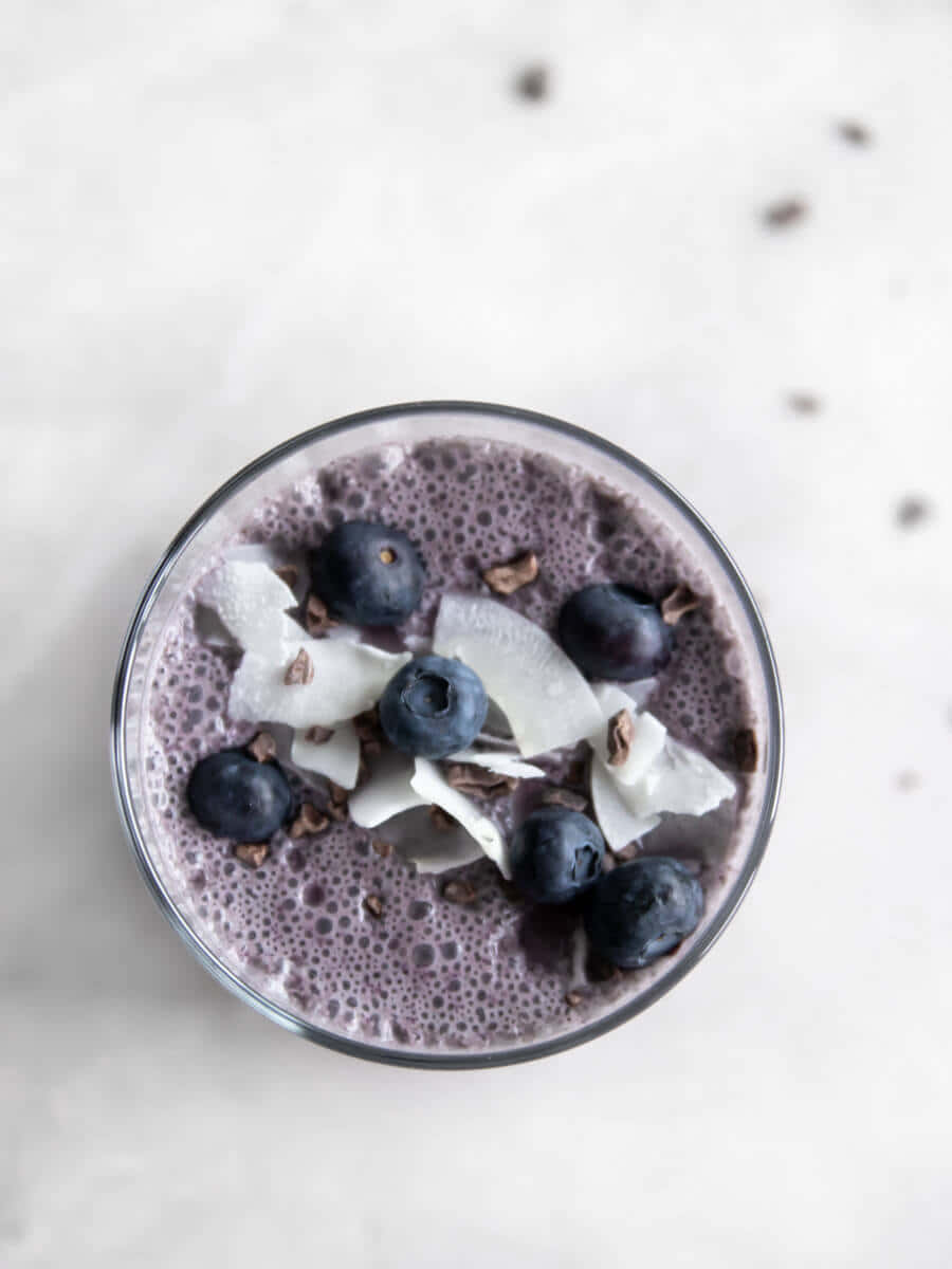 Blueberry Smoothie 桌布 壁紙 — 一個俯視角度，顯示一杯紫色冰沙，上面放有藍莓和椰子片，置於淺色斑點表面上。