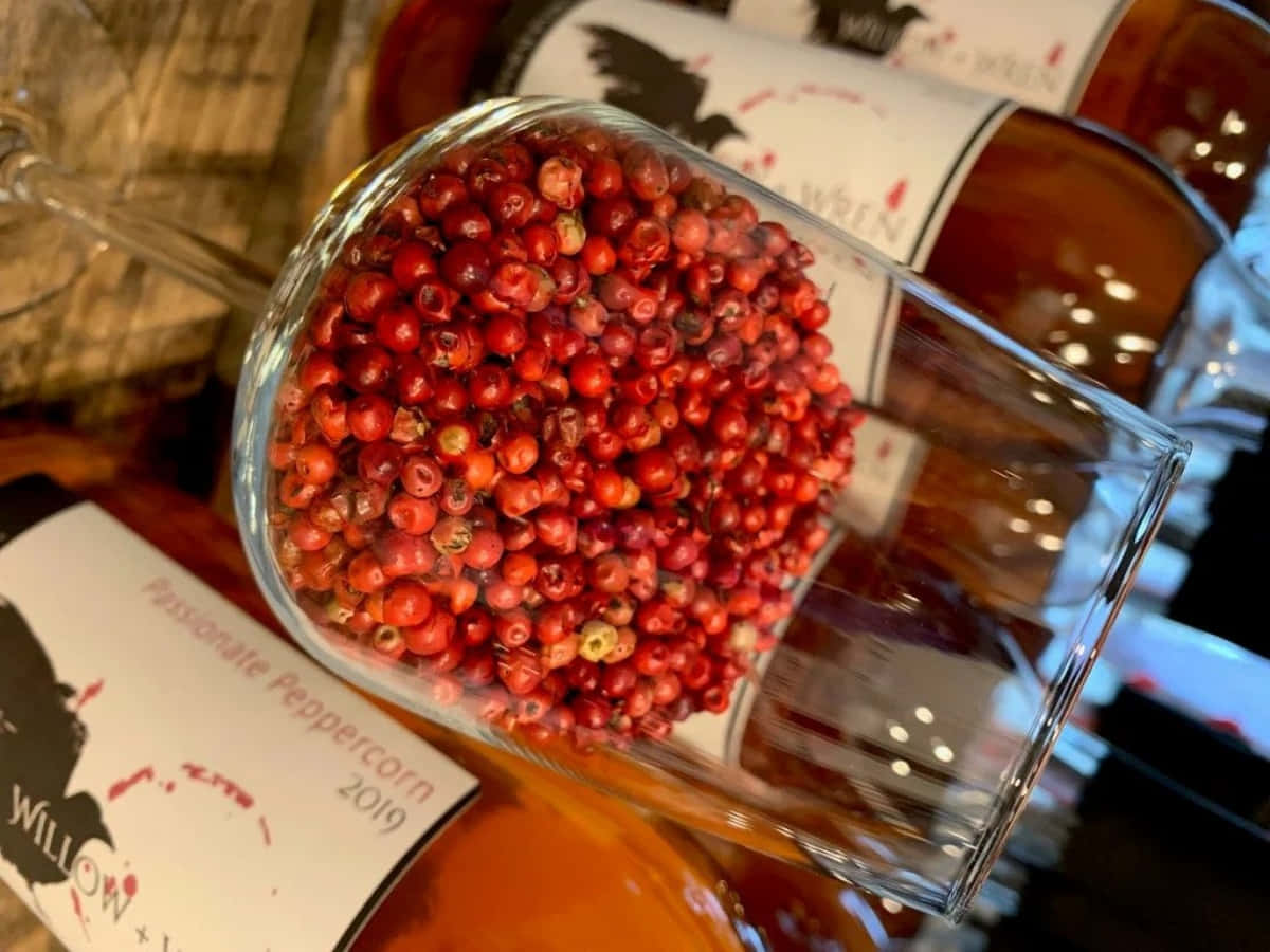 Pink Peppercorn 桌布 壁紙 — 一個傾斜的玻璃杯盛滿鮮紅的胡椒粒，放在標有「Passionate Peppercorn 2019」的瓶子上，環境溫暖明亮。