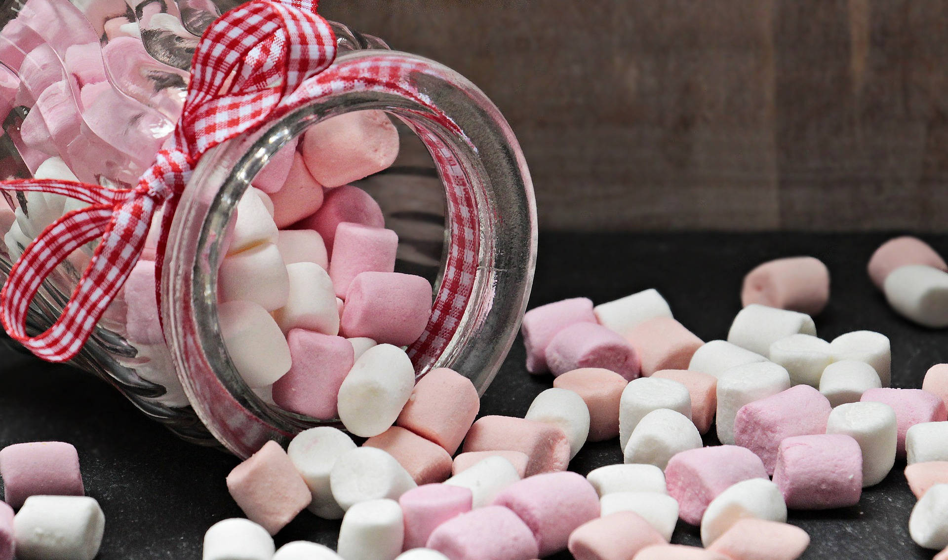 Marshmallow 桌布 壁紙 — 一個傾斜的玻璃罐，帶有紅白格子蝴蝶結，將粉紅色和白色的棉花糖灑在深色石板表面上。