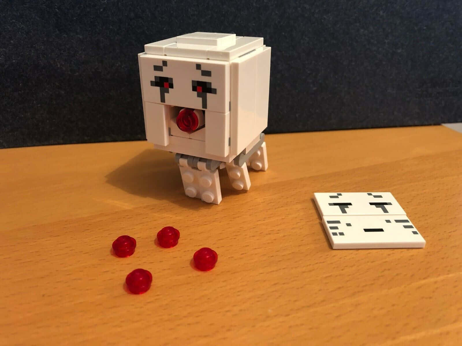 Minecraft Ghast 桌布 壁紙 — 一個像素化的白色LEGO風格生物，有紅色眼睛和嘴巴，坐在木桌上，旁邊散落著紅色珠子和一個分離的面板。
