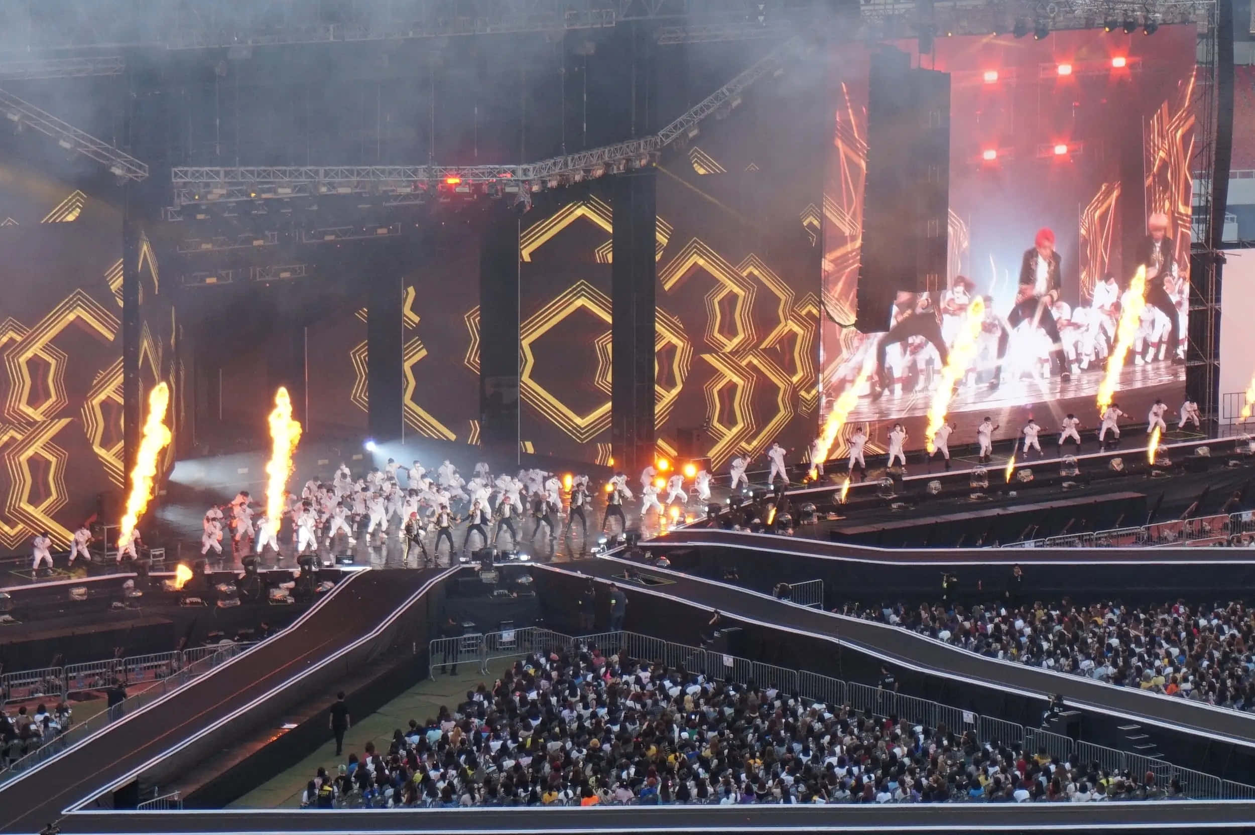 Bts Stage 桌布 壁紙 — 一個充滿動感的演唱會舞台，表演者身穿白色服裝，在熾熱的煙火和發光的幾何屏幕中跳舞。