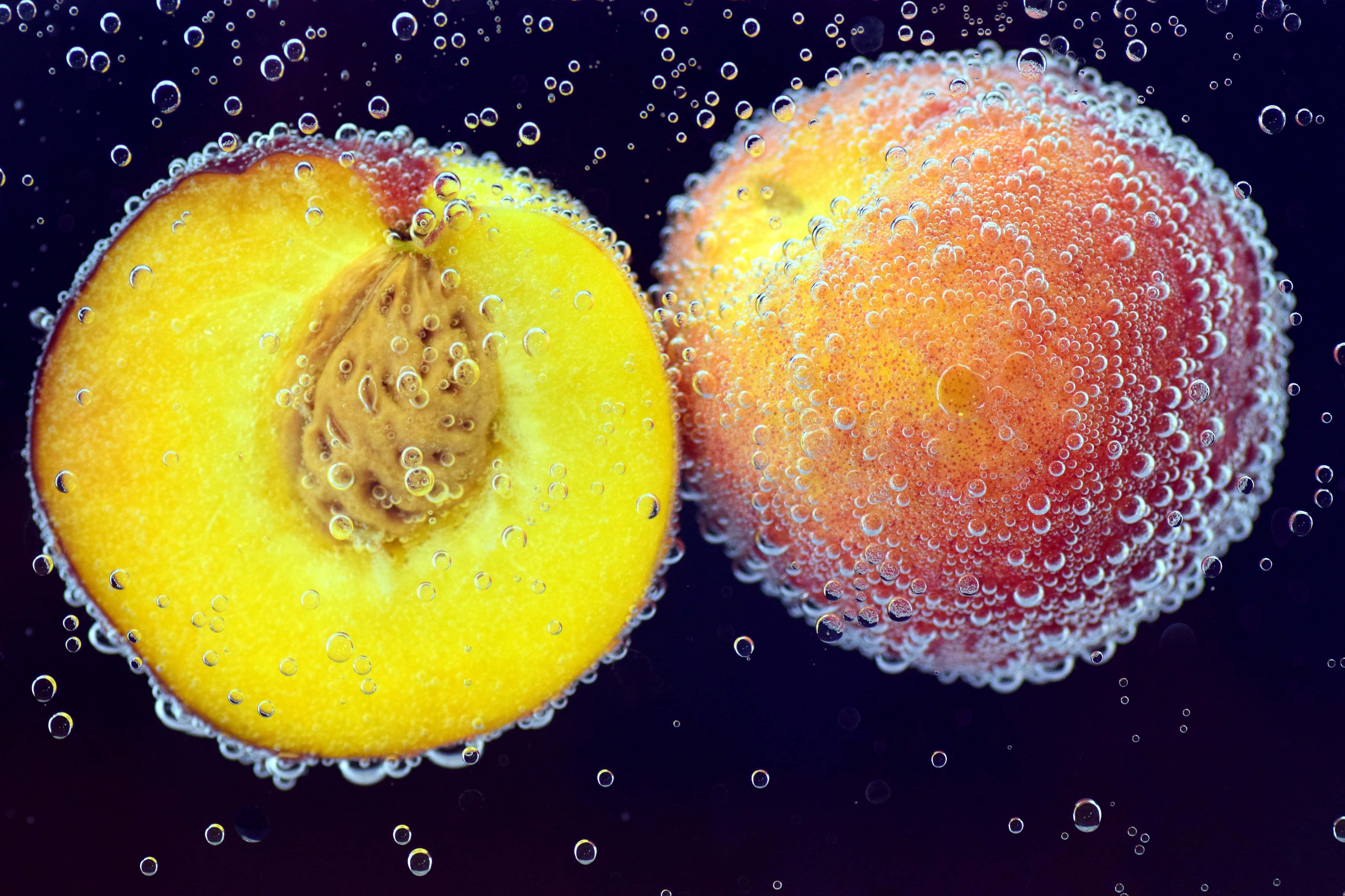 Summer Fruits 桌布 壁紙 — 一個充滿活力的半個桃子和整個桃子浸在深色液體中，周圍有閃閃發光的氣泡。