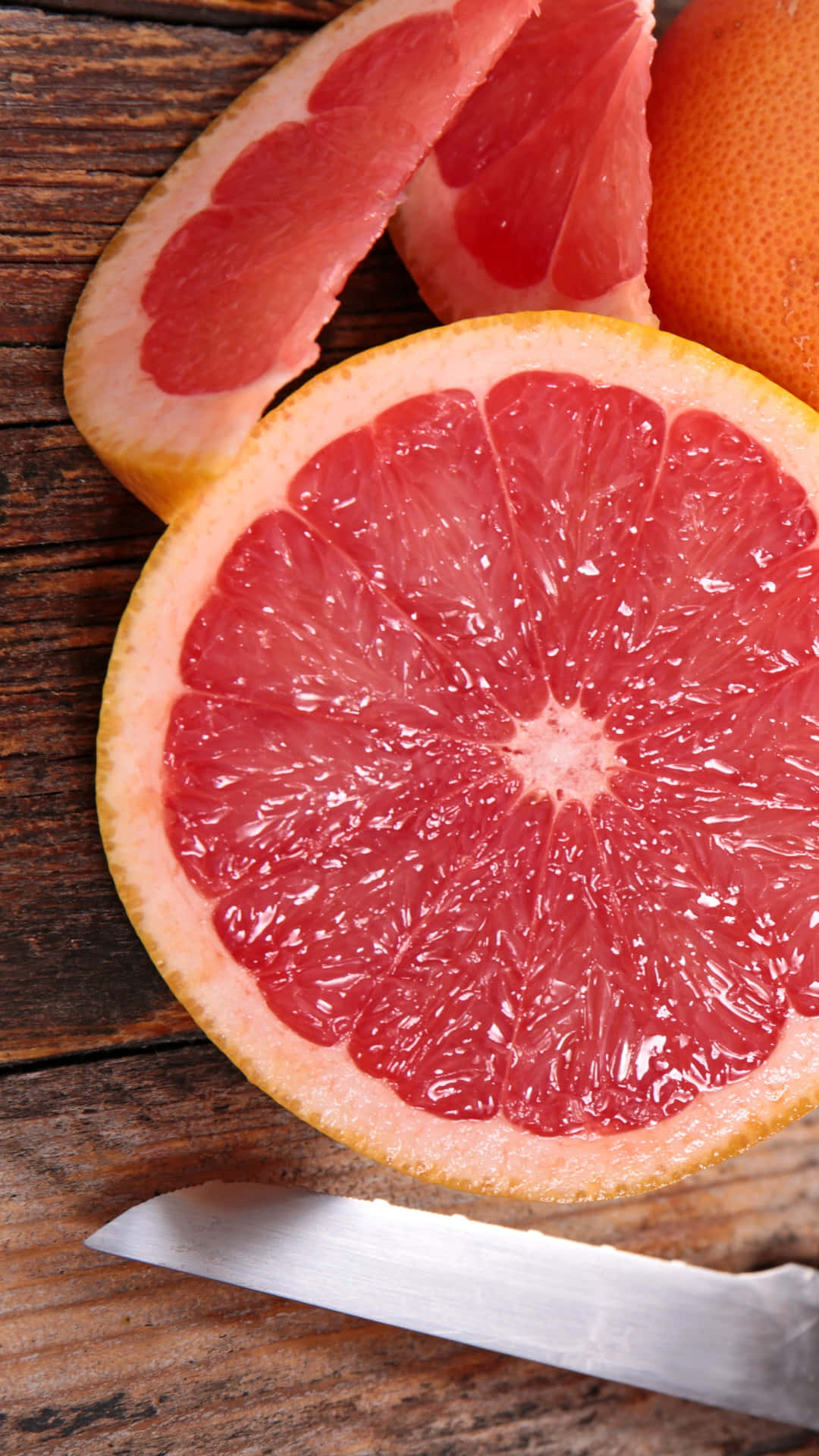 Pink Grapefruit 桌布 壁紙 — 一個充滿活力的半切西柚，帶有多汁的粉紅色果肉，放在質樸的木桌上，旁邊有一把刀和完整的水果。