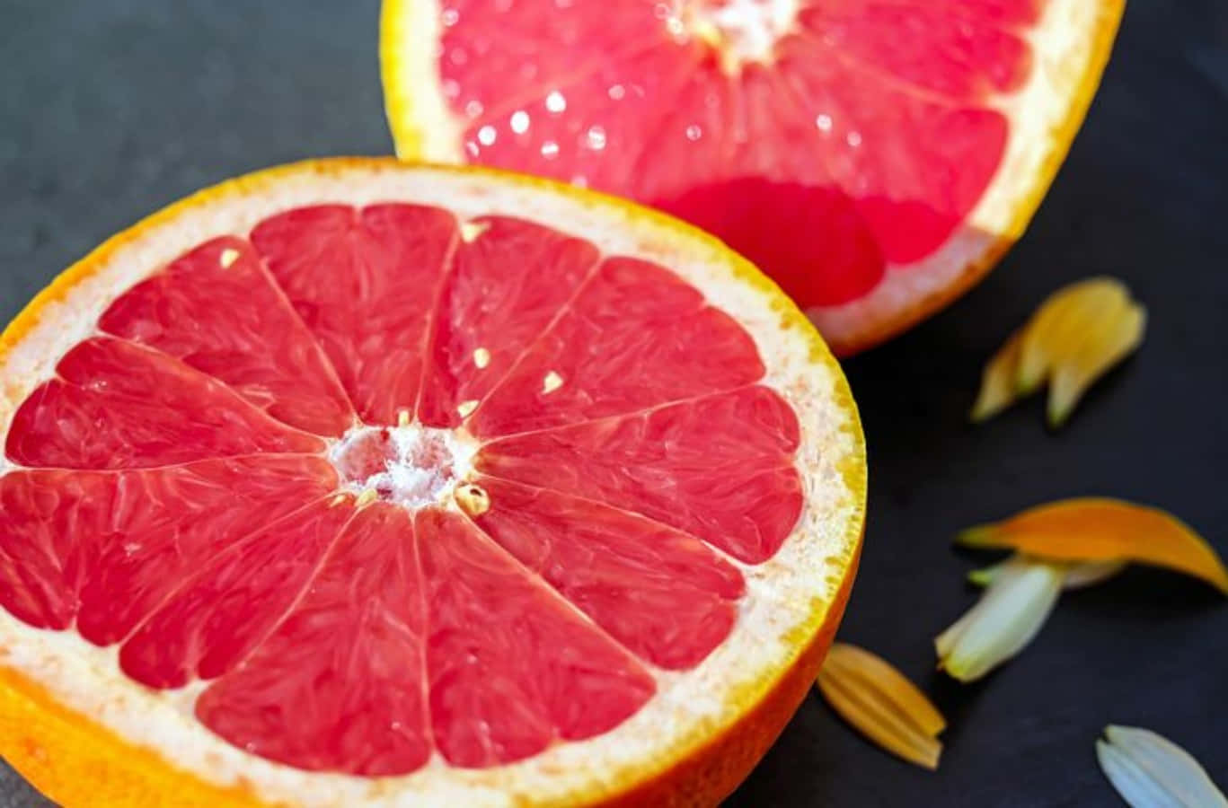 Pink Grapefruit 桌布 壁紙 — 一個充滿活力的半切西柚在深色石板表面上露出多汁的粉紅色果肉，周圍散落著花瓣。