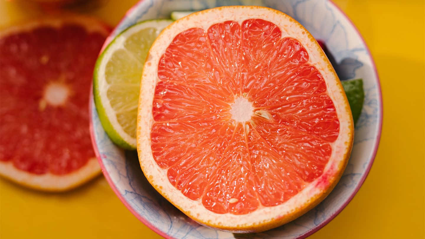 Winter Fruits 桌布 壁紙 — 一個充滿活力的半切西柚配青檸片，放在一個有圖案的碗裡，背景是明亮的黃色。