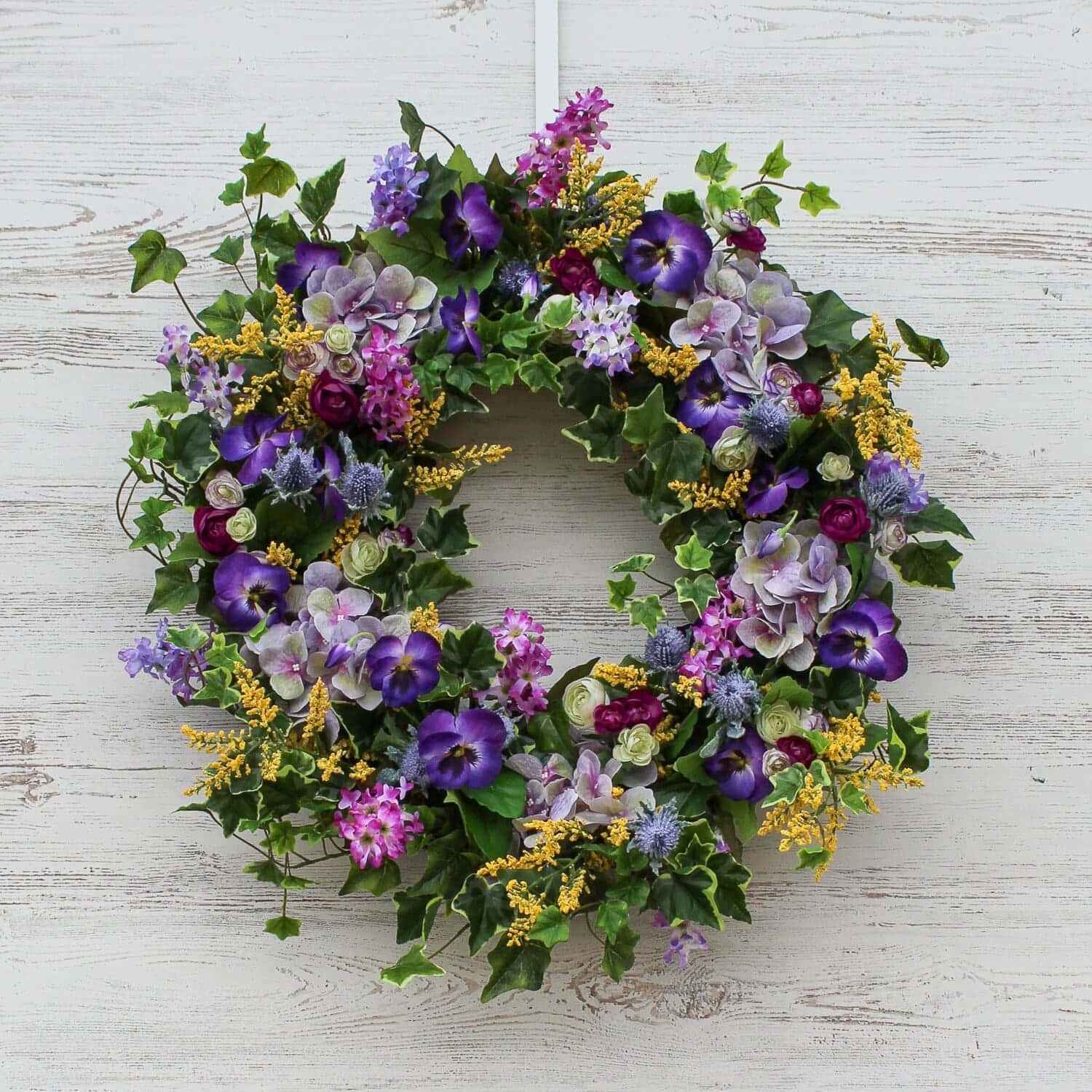 Spring Wreaths 桌布 壁紙 — 一個充滿活力的圓形花環，飾有紫色、粉紅色和黃色的花朵以及綠色常春藤，掛在風化的白色木牆上。