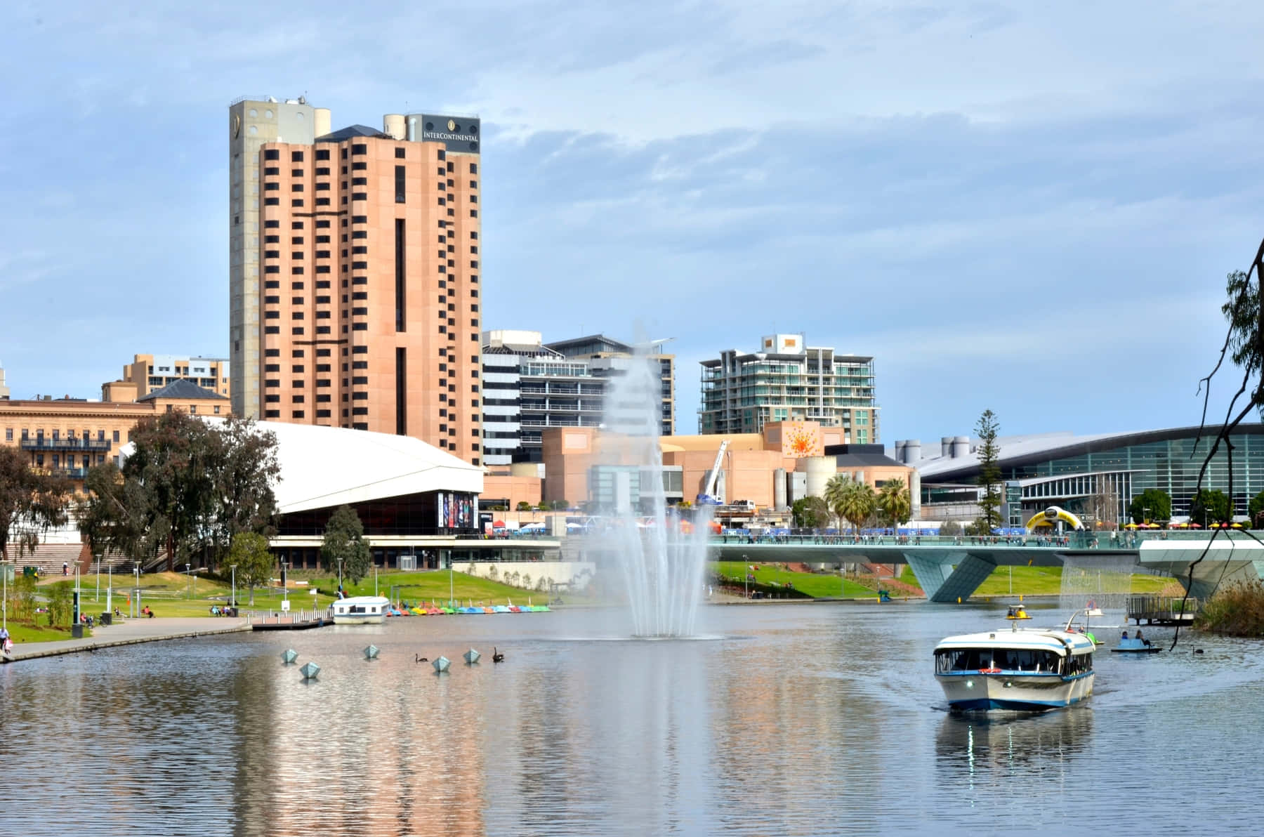Adelaide 桌布 壁紙 — 一個充滿活力的城市海濱場景，有一座高磚酒店、中央噴泉和船隻在部分多雲的天空下平靜的水面上滑行。