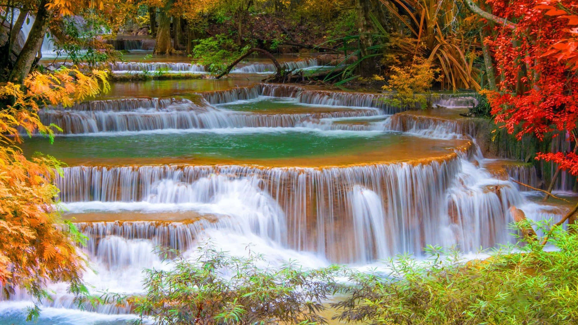 Waterfalls 桌布 壁紙 — 一個充滿活力的多層瀑布穿過秋葉傾瀉而下，火紅、橙和綠色的樹葉框住了藍綠色的水。