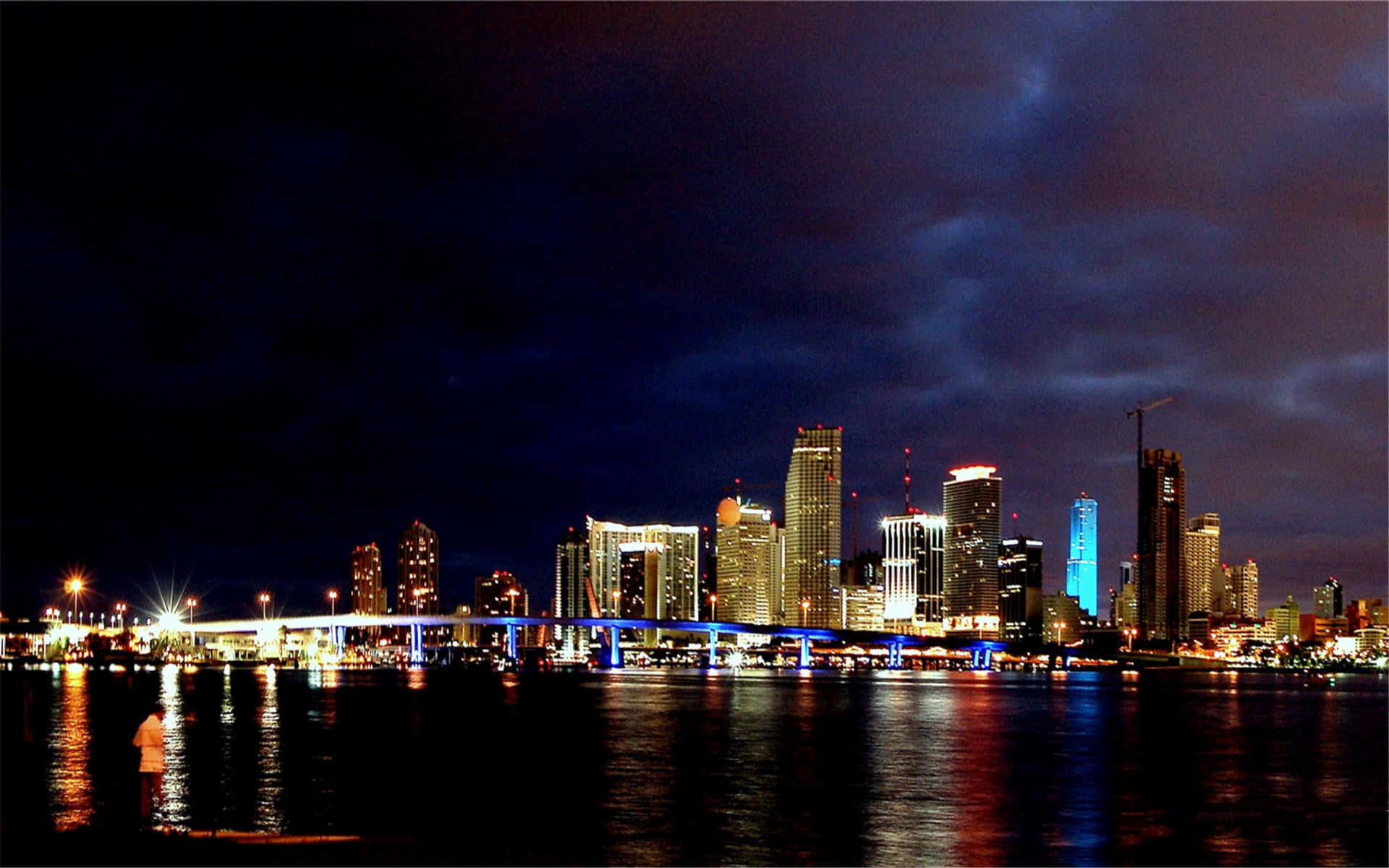 邁阿密 桌布 壁紙 — 一個充滿活力的夜景城市景觀，在陰天之下，漆黑的湖面倒映著燈火通明的摩天大樓和一座發光的橋樑。
