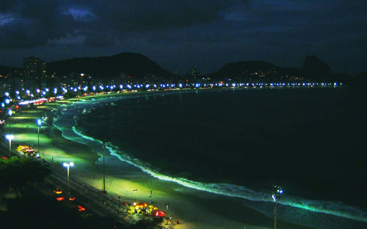 Brazil 桌布 壁紙 — 一個充滿活力的夜間海灘場景，有發光的藍綠色海浪、燈火通明的木板路，以及遠處城市燈光映襯著黑暗的山丘。