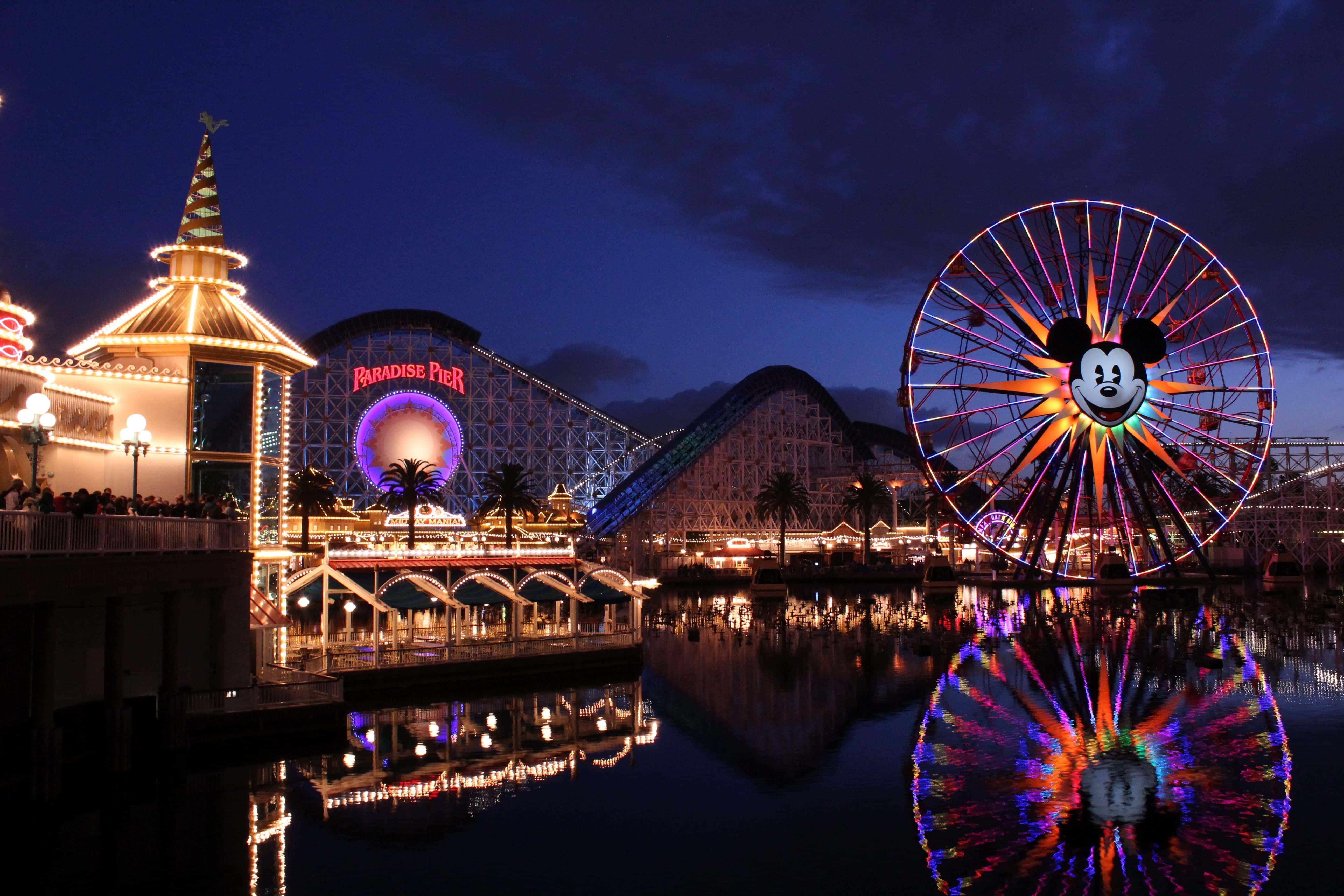 Disneyland 桌布 壁紙 — 一個充滿活力的夜間遊樂園，發光的米奇老鼠摩天輪和過山車倒映在平靜的水面上。