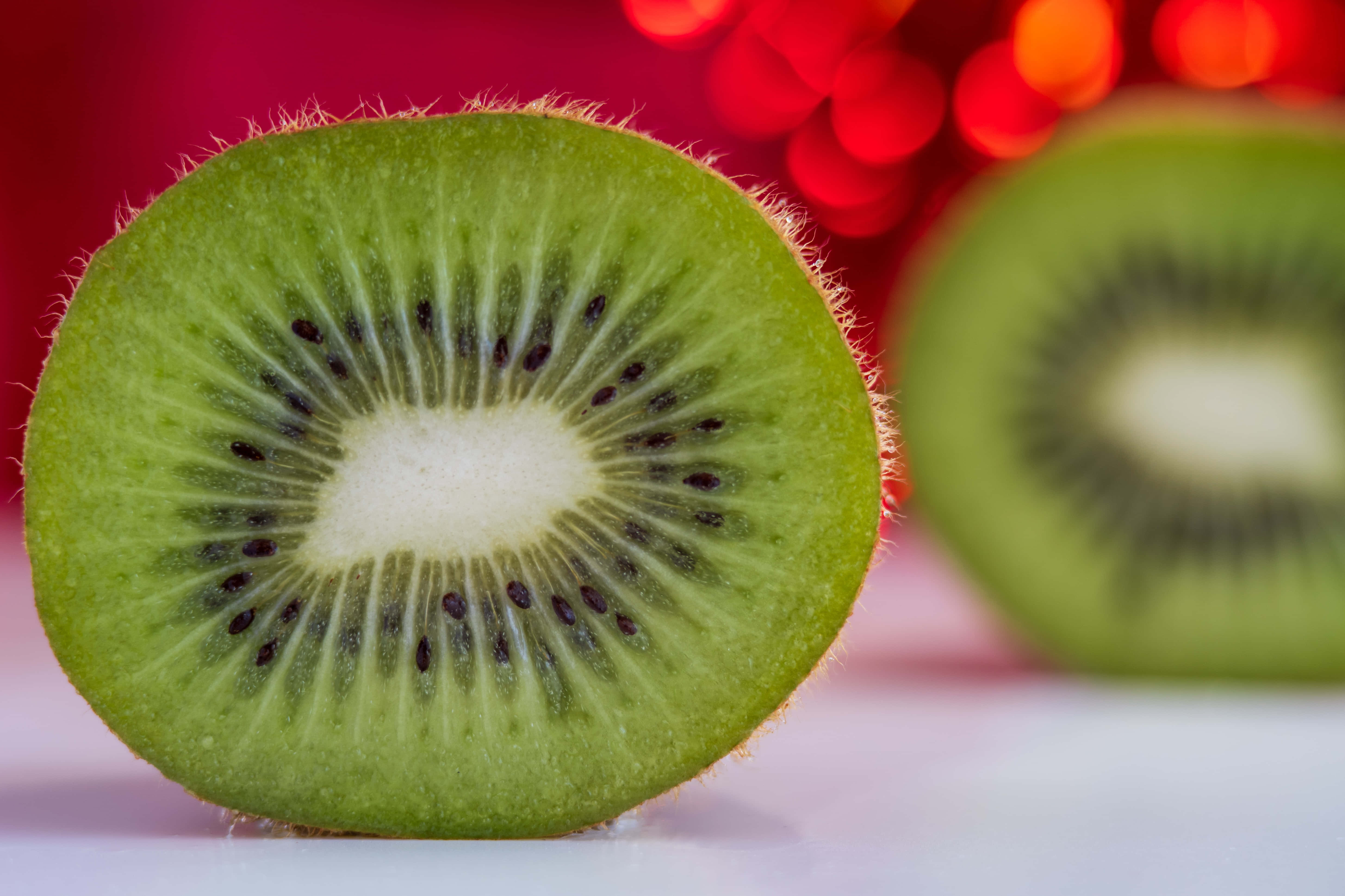 Kiwi 桌布 壁紙 — 一個充滿活力的奇異果切片特寫，帶有毛茸茸的綠色外皮和黑色種子，背景是模糊的紅色散景。
