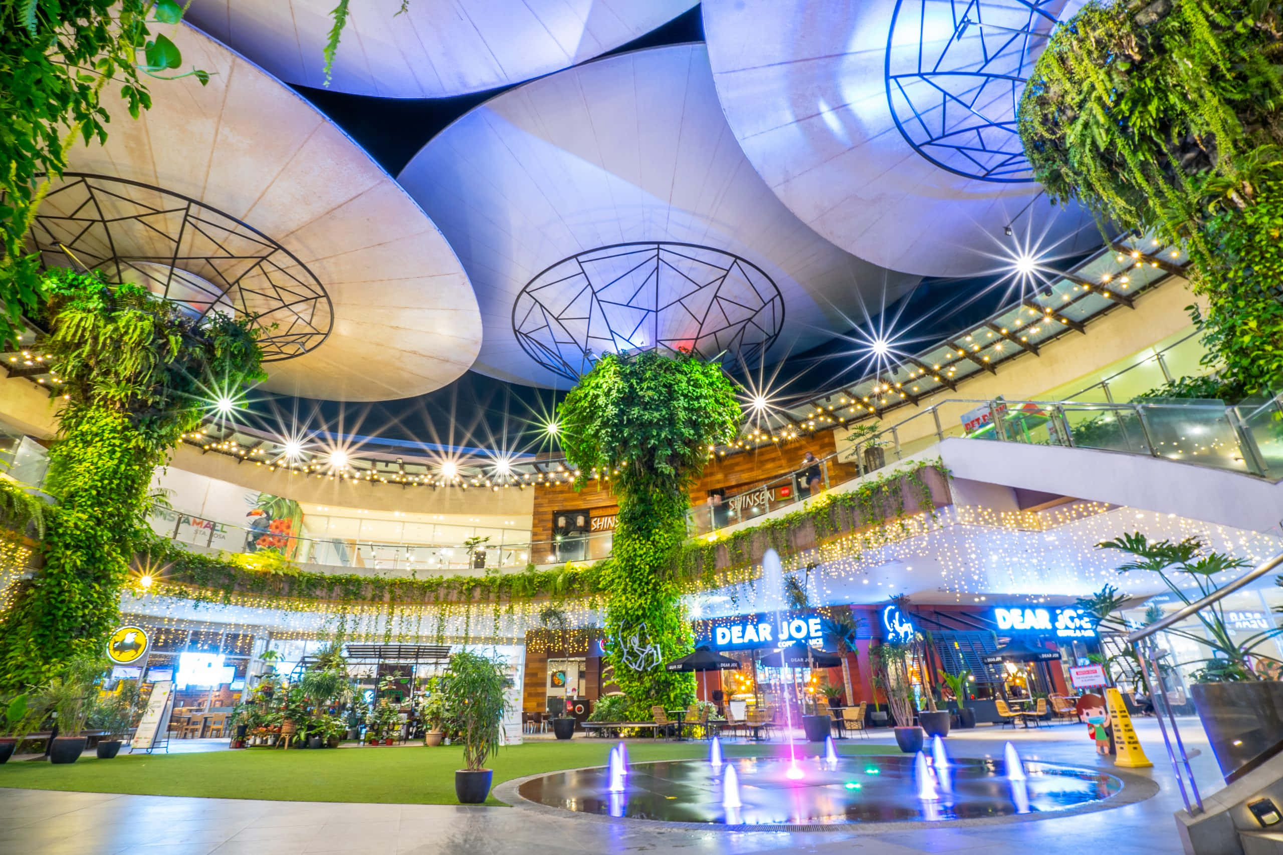 Mall 桌布 壁紙 — 一個充滿活力的室內購物中心中庭，帶有從未來感弧形天花板傾瀉而下的發光燈光，以及攀爬在柱子上的茂密綠色植物。
