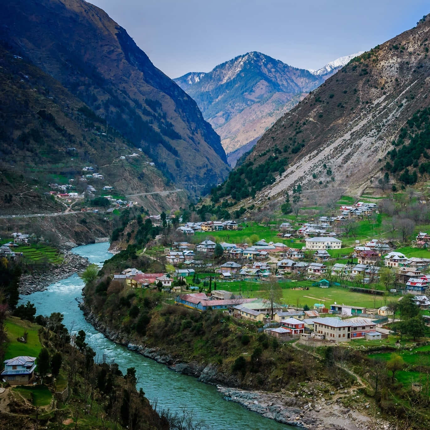 Pakistan 桌布 壁紙 — 一個充滿活力的山村坐落在山谷中，一條湍急的碧綠河流蜿蜒穿過茂密的綠色田野和陡峭的岩石斜坡。
