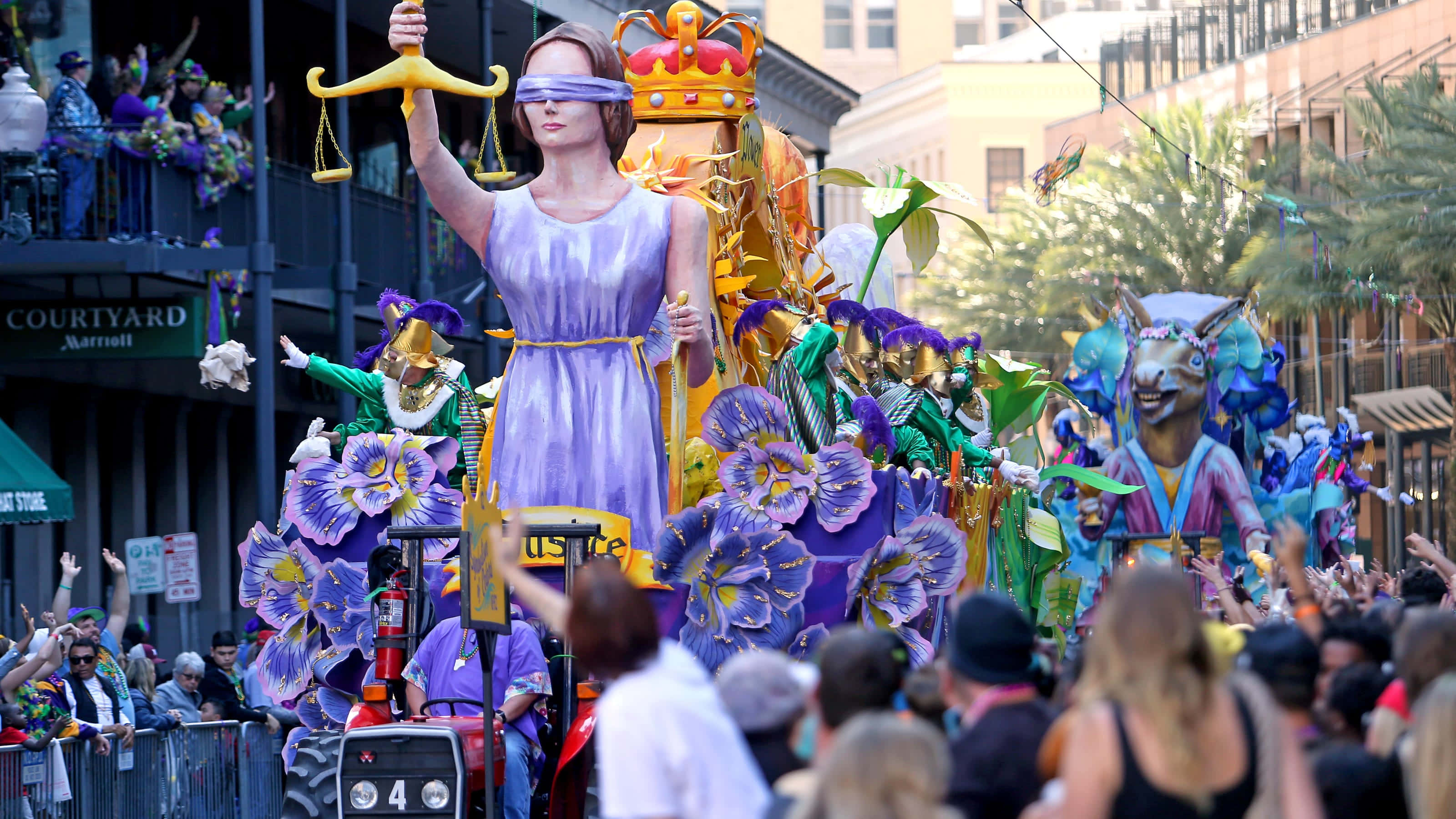 Mardi Gras 桌布 壁紙 — 一個充滿活力的巡遊花車，展示了一位蒙眼的正義女神，手持天平，周圍是色彩繽紛的花卉裝飾和歡呼的人群。