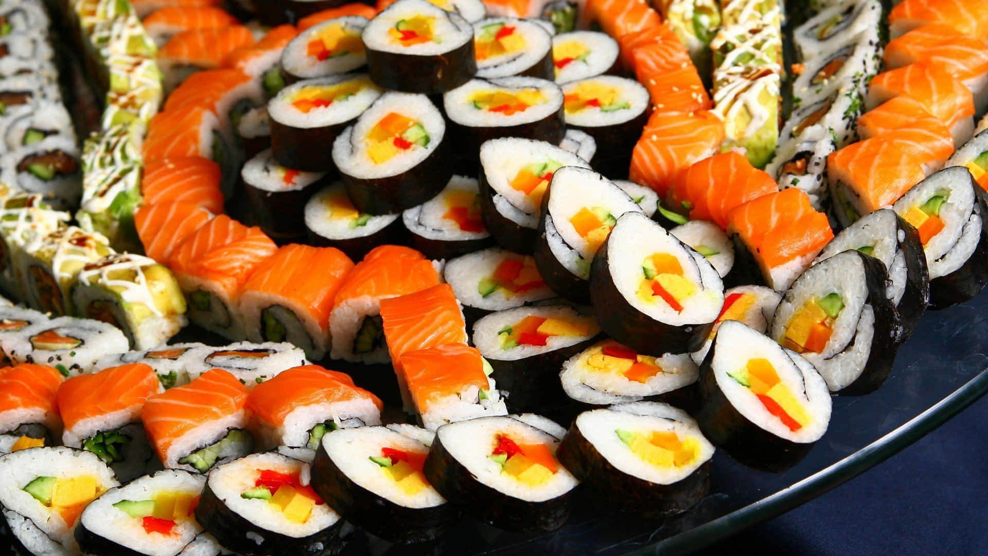 Sushi 桌布 壁紙 — 一個充滿活力的拼盤，包含各式壽司卷和刺身，以色彩繽紛、誘人的圖案排列。
