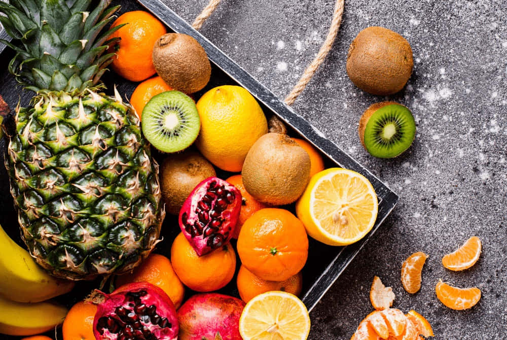 Winter Fruits 桌布 壁紙 — 一個充滿活力的新鮮熱帶和柑橘類水果組合，如菠蘿、奇異果、石榴和橙子，排列在一個質樸的黑色托盤上，置於有紋理的灰色表面。