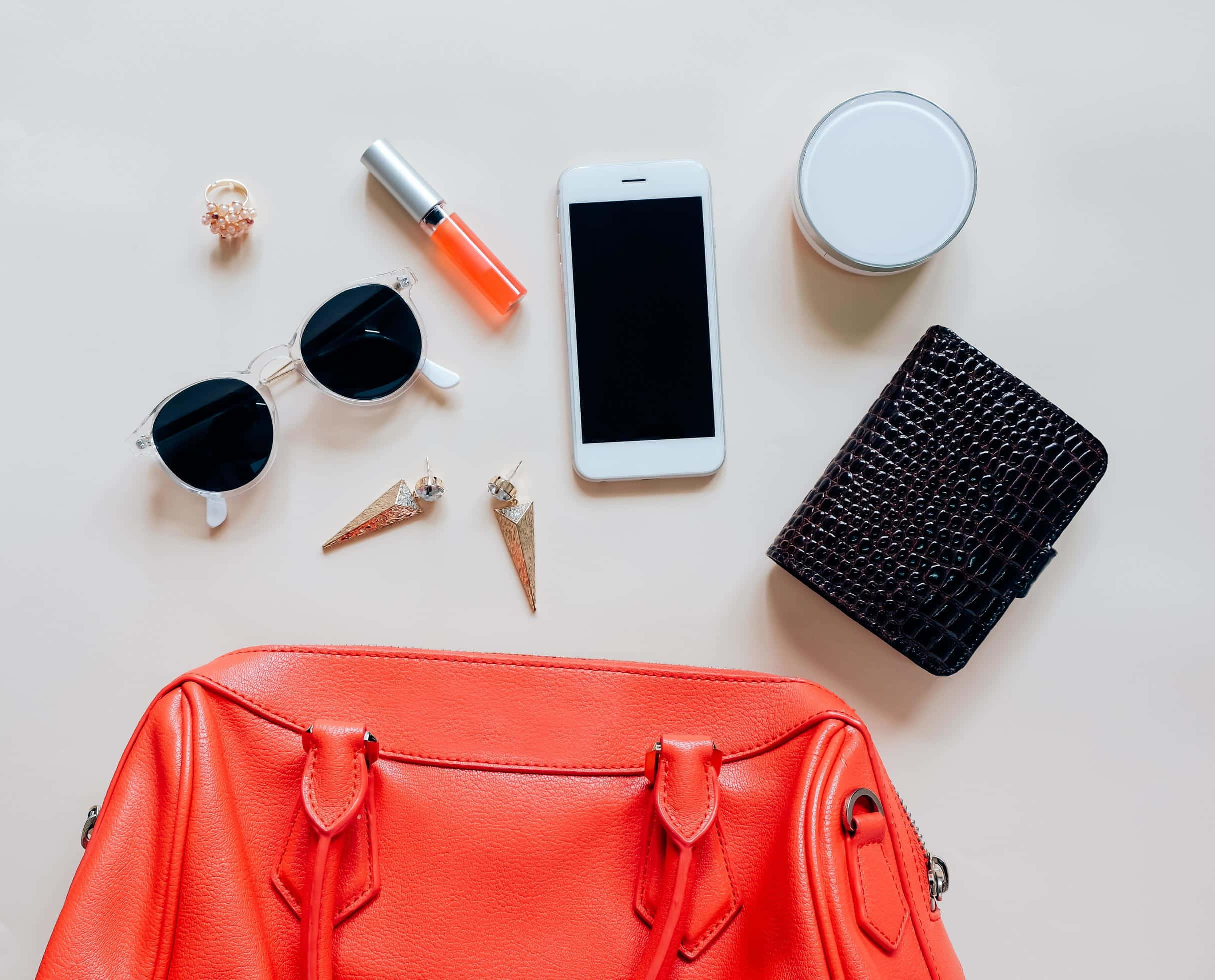 Red Handbag 桌布 壁紙 — 一個充滿活力的橙色皮革手袋放在中央，太陽眼鏡、唇膏、電話和配件整齊地排列在淺米色表面上。