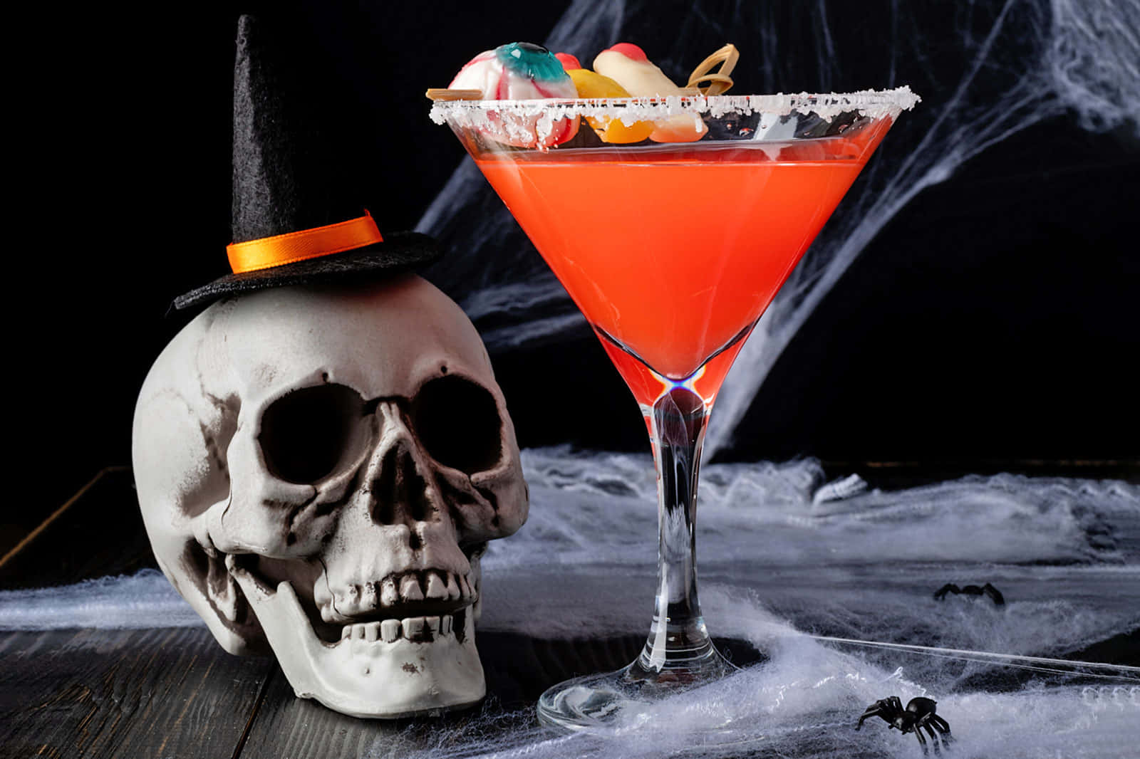 Halloween Cocktails 桌布 壁紙 — 一個充滿活力的橙色萬聖節雞尾酒放在馬提尼杯中，旁邊是一個戴著巫婆帽的骷髏頭，放在佈滿蜘蛛網的桌子上。