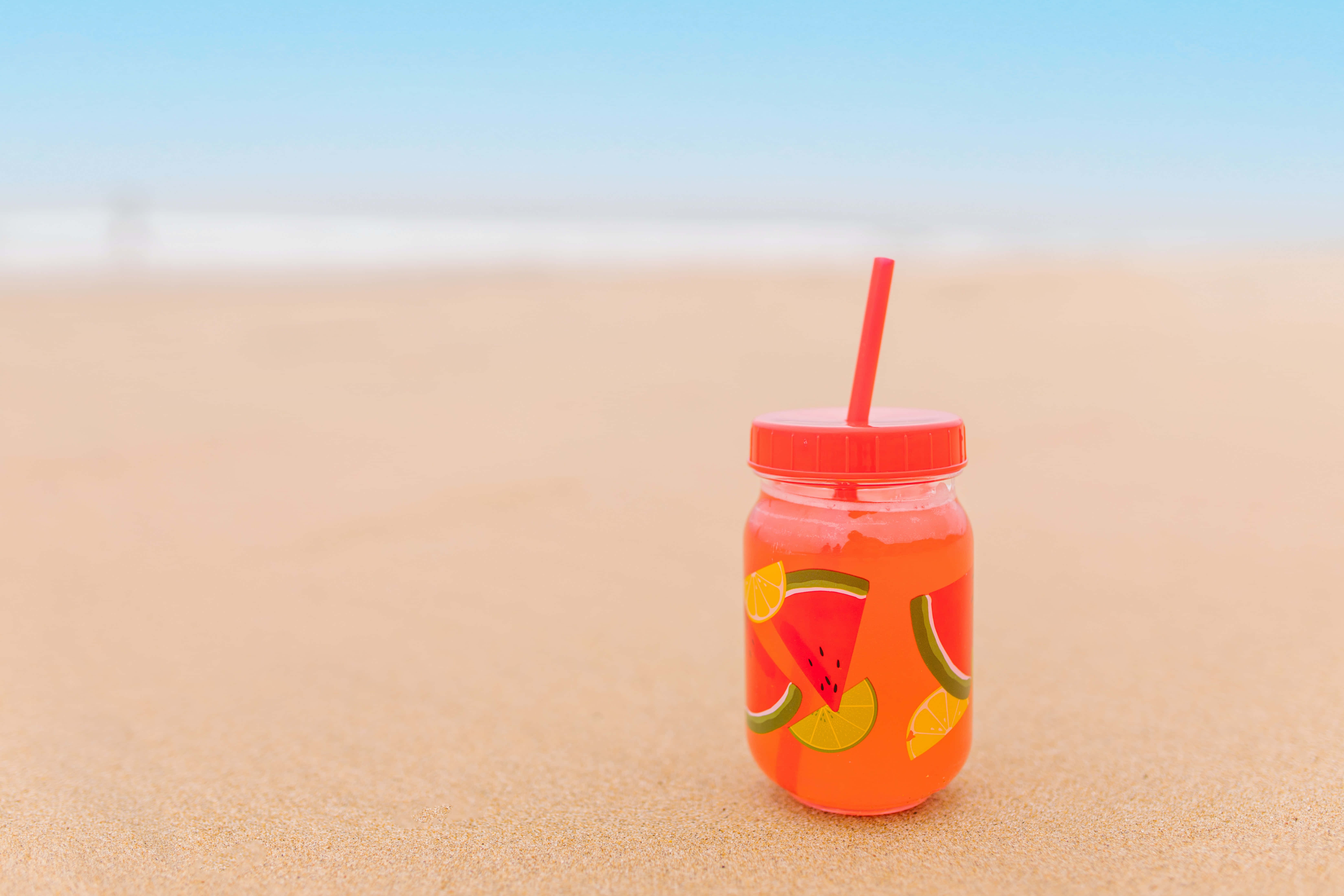 Summer Drinks 桌布 壁紙 — 一個充滿活力的橙色西瓜汁瓶，配紅色飲管，放在金色沙灘上，天空湛藍清澈。