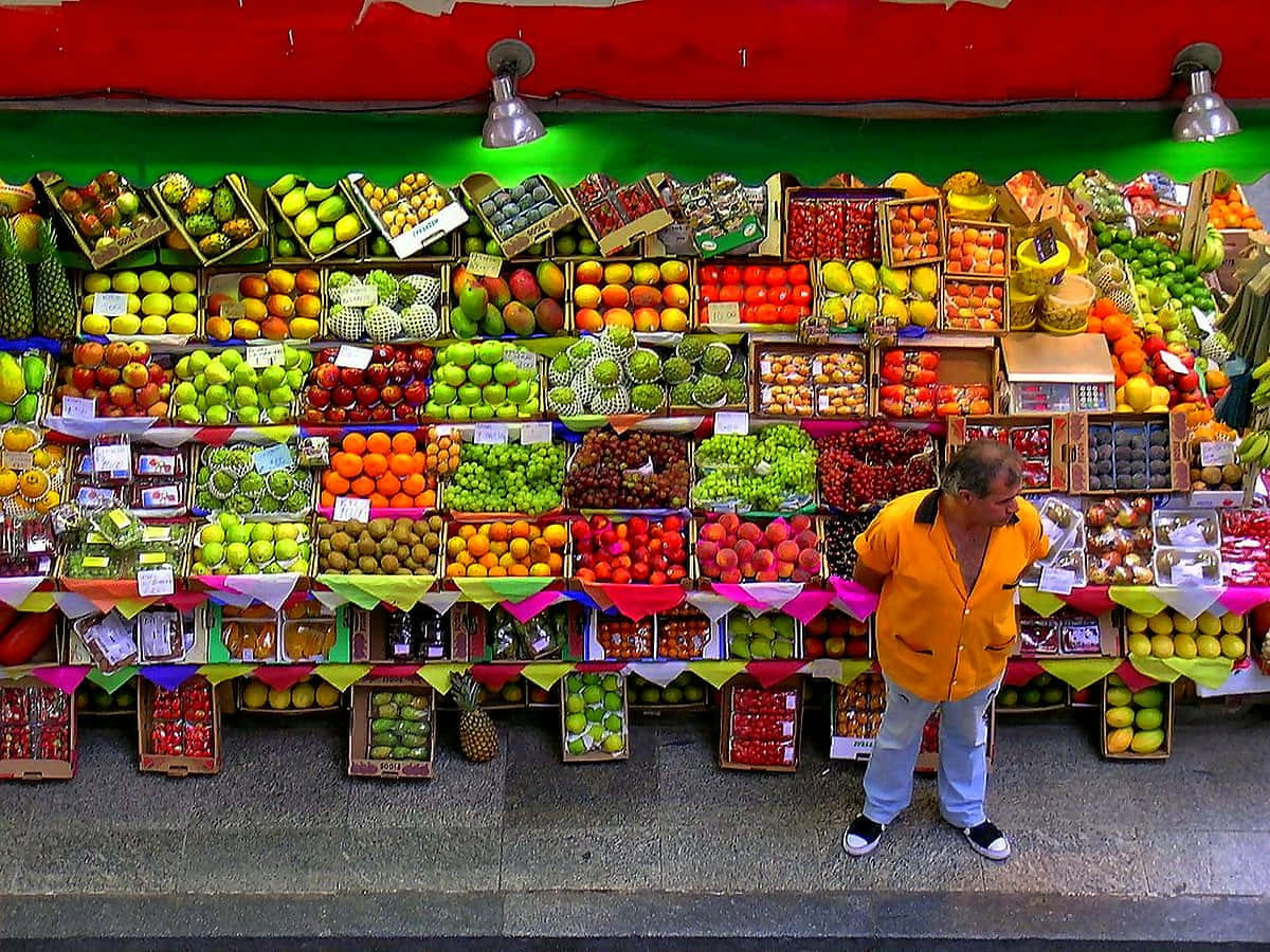 Grocery Store 桌布 壁紙 — 一個充滿活力的水果攤在明亮的燈光下擺滿了色彩繽紛的產品，一名身穿橙色襯衫的男子正在瀏覽商品。