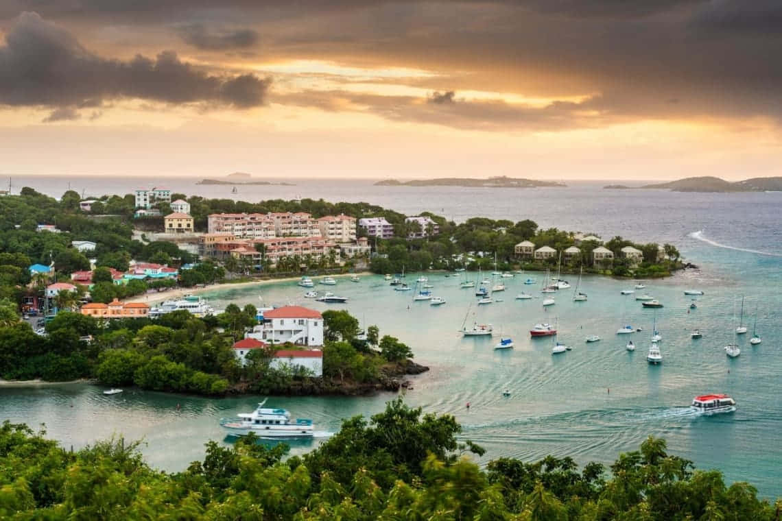 Caribbean Island 桌布 壁紙 — 一個充滿活力的沿海小鎮，坐落在鬱鬱蔥蔥的海灣中，日落時分，戲劇性的雲層下，船隻點綴在碧綠的海水上。