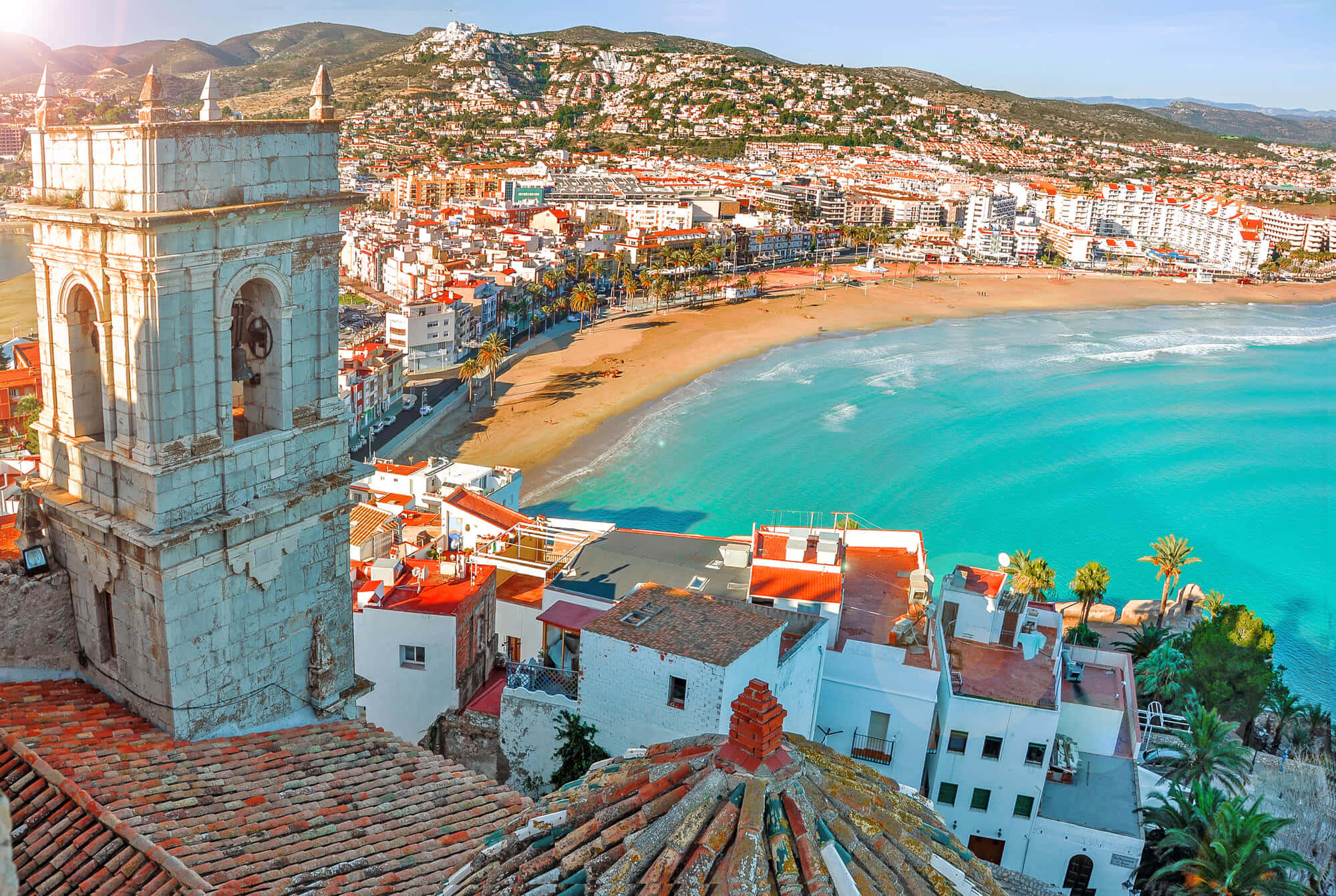 Spanish Beach 桌布 壁紙 — 一個充滿活力的沿海小鎮，白色建築和紅色屋頂環抱著一個碧綠的海灣，上方是清澈的藍天。