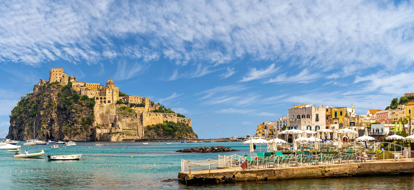 Italian Island 桌布 壁紙 — 一個充滿活力的沿海小鎮，色彩繽紛的建築物依偎在海濱碼頭旁，而一座歷史悠久的堡壘則矗立在岩石島上，天空湛藍，雲朵輕柔。