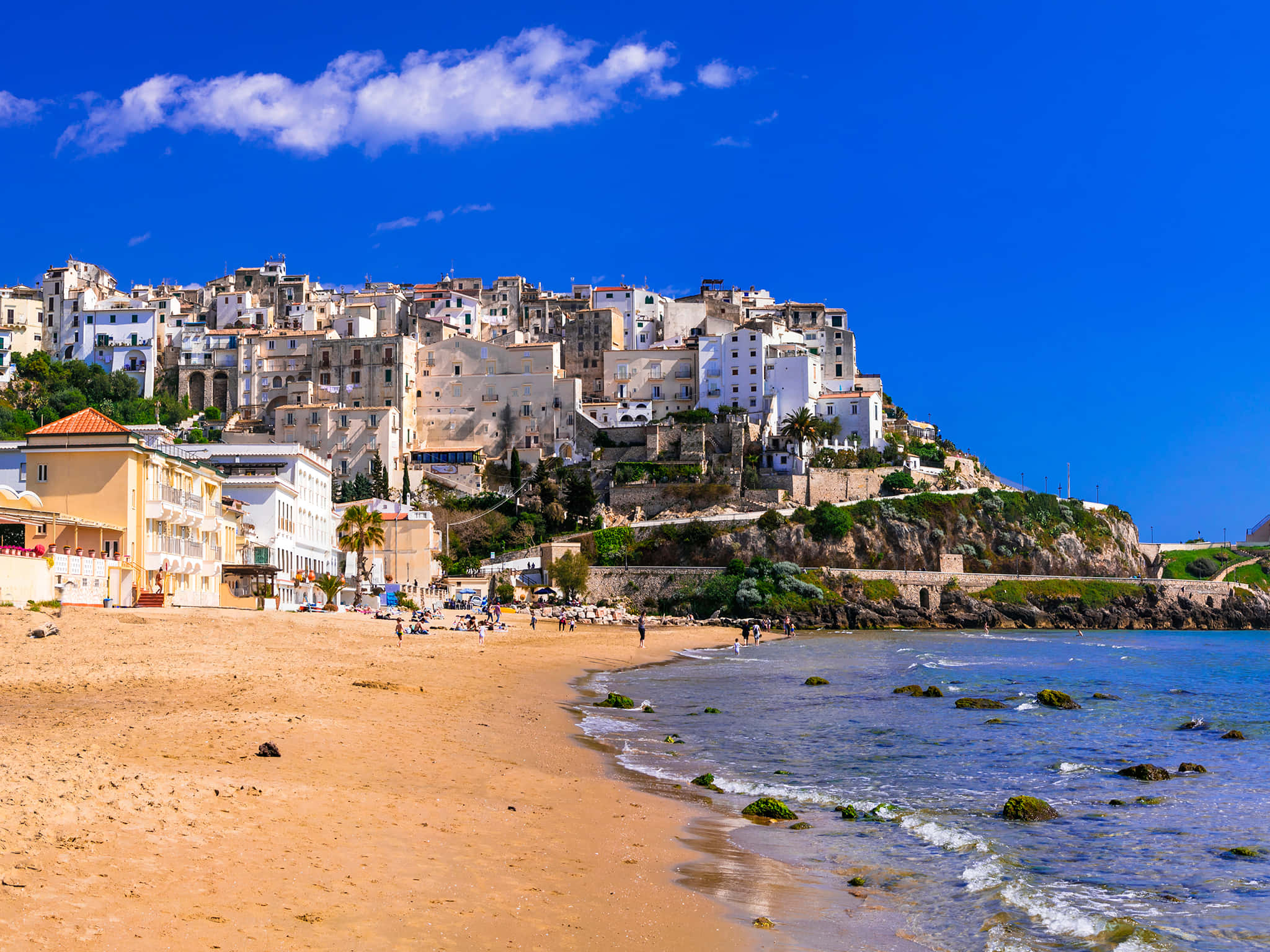 Italian Beach 桌布 壁紙 — 一個充滿活力的沿海小鎮依附在陽光普照的懸崖上，俯瞰著金色的沙灘和碧綠的海水，天空湛藍明亮。