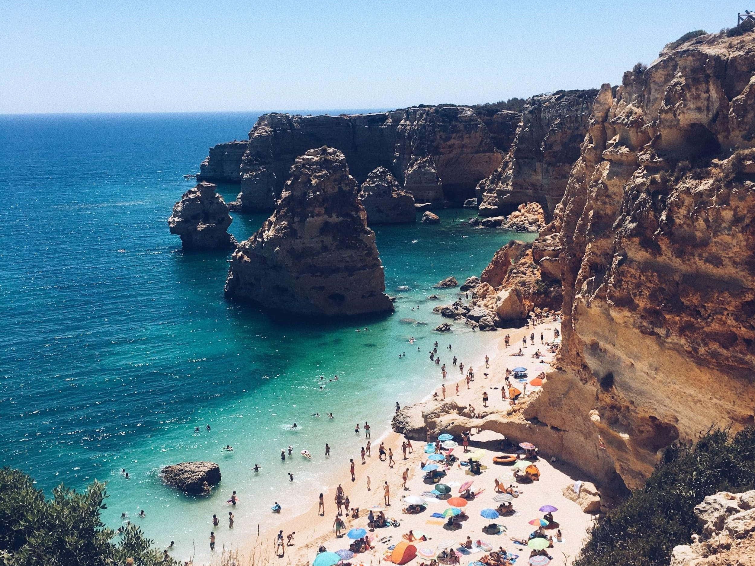 Portuguese Beach 桌布 壁紙 — 一個充滿活力的海灘，坐落於高聳的金色懸崖之間，擁有碧綠的海水和色彩繽紛的太陽傘下的日光浴者。