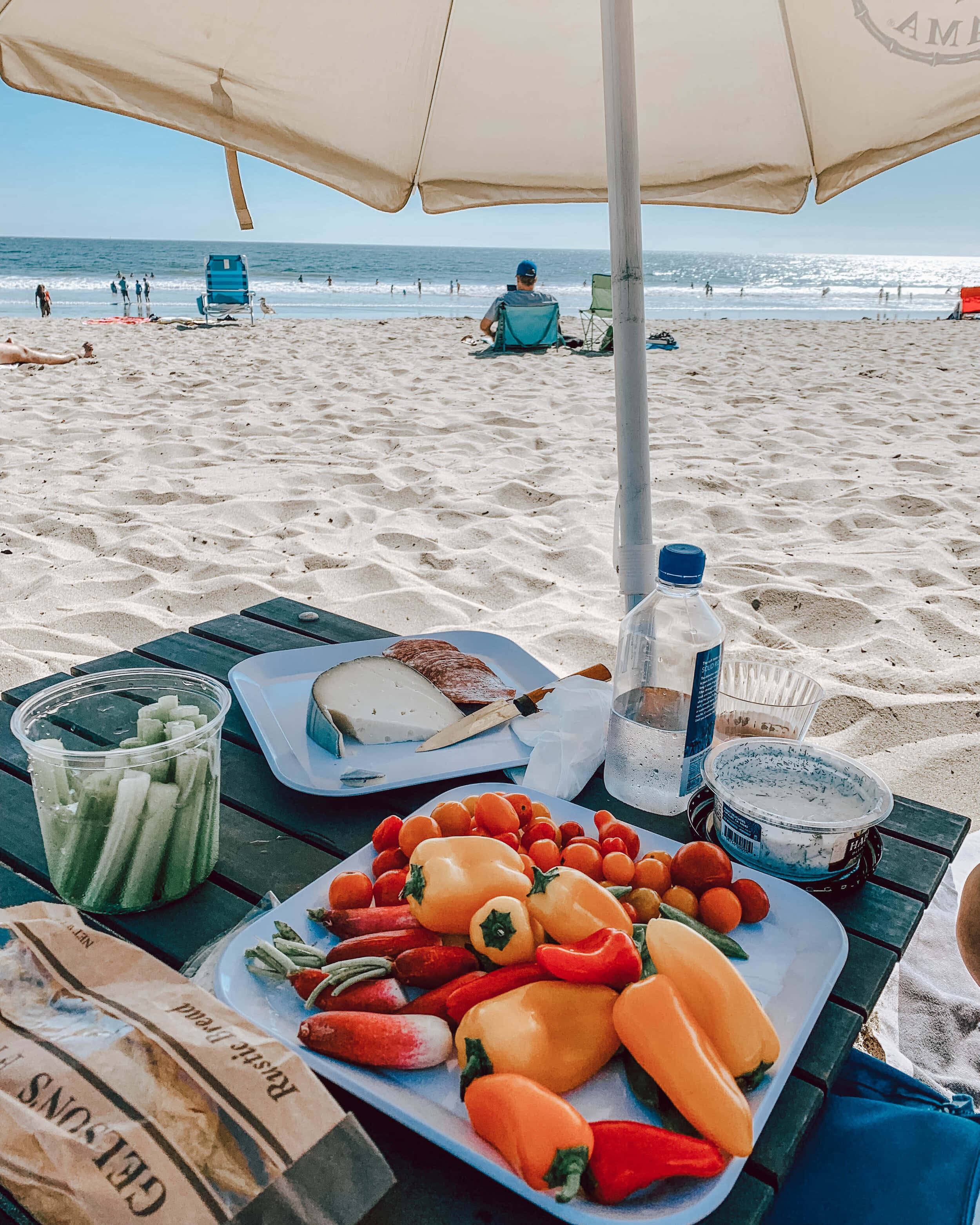 Beach Picnic 桌布 壁紙 — 一個充滿活力的海灘野餐，有色彩繽紛的燈籠椒、番茄和麵包，放在米色遮陽傘下，俯瞰海洋。