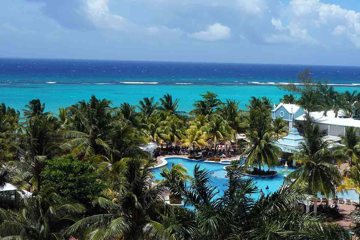 Jamaican Beach 桌布 壁紙 — 一個充滿活力的熱帶度假勝地，擁有綠松石色的泳池和棕櫚樹，俯瞰著部分多雲天空下壯麗的藍色海洋。