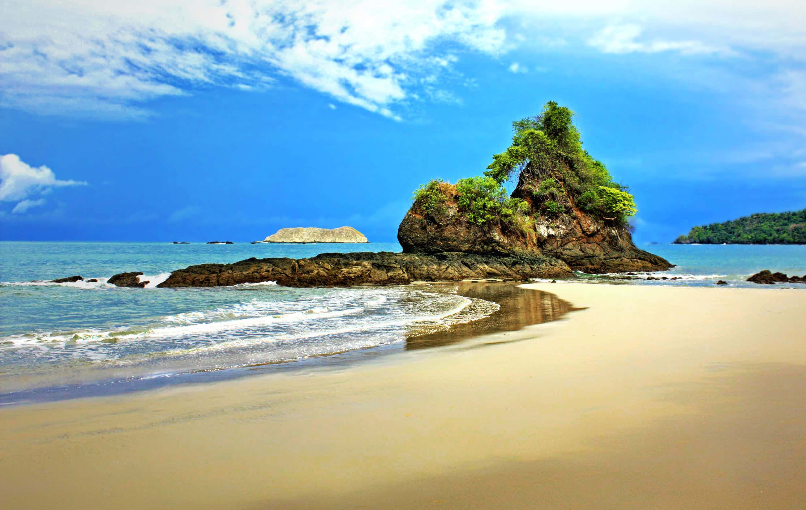 Costa Rica 桌布 壁紙 — 一個充滿活力的熱帶海灘，有金色沙灘、溫柔的海浪，以及從碧綠海水中升起、在明亮藍天下茂密的綠色岩石地貌。