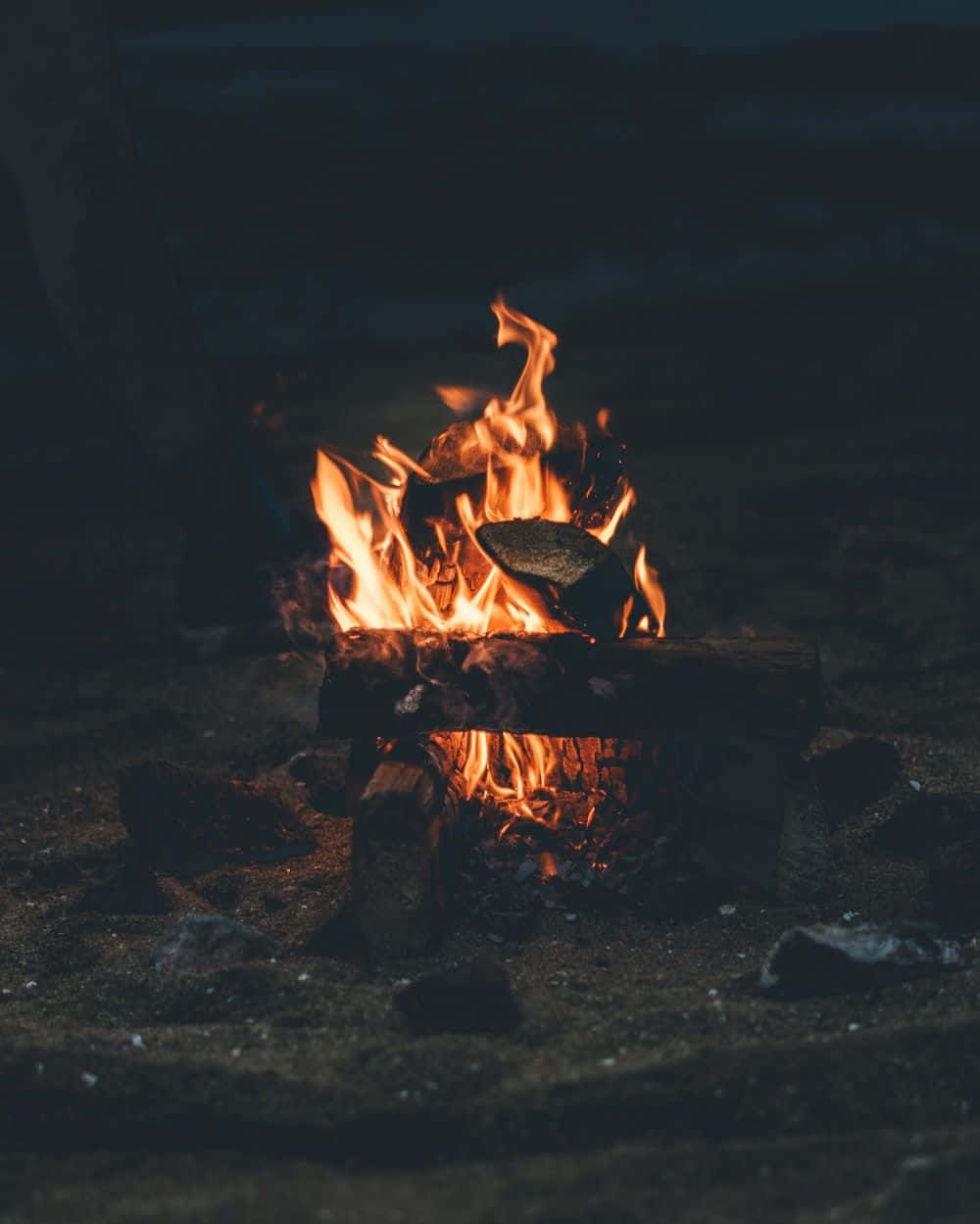 Beach Camping 桌布 壁紙 — 一個充滿活力的營火在漆黑的夜空背景下噼啪作響，橙色的火焰將溫暖的光芒投射在周圍的岩石和泥土上。