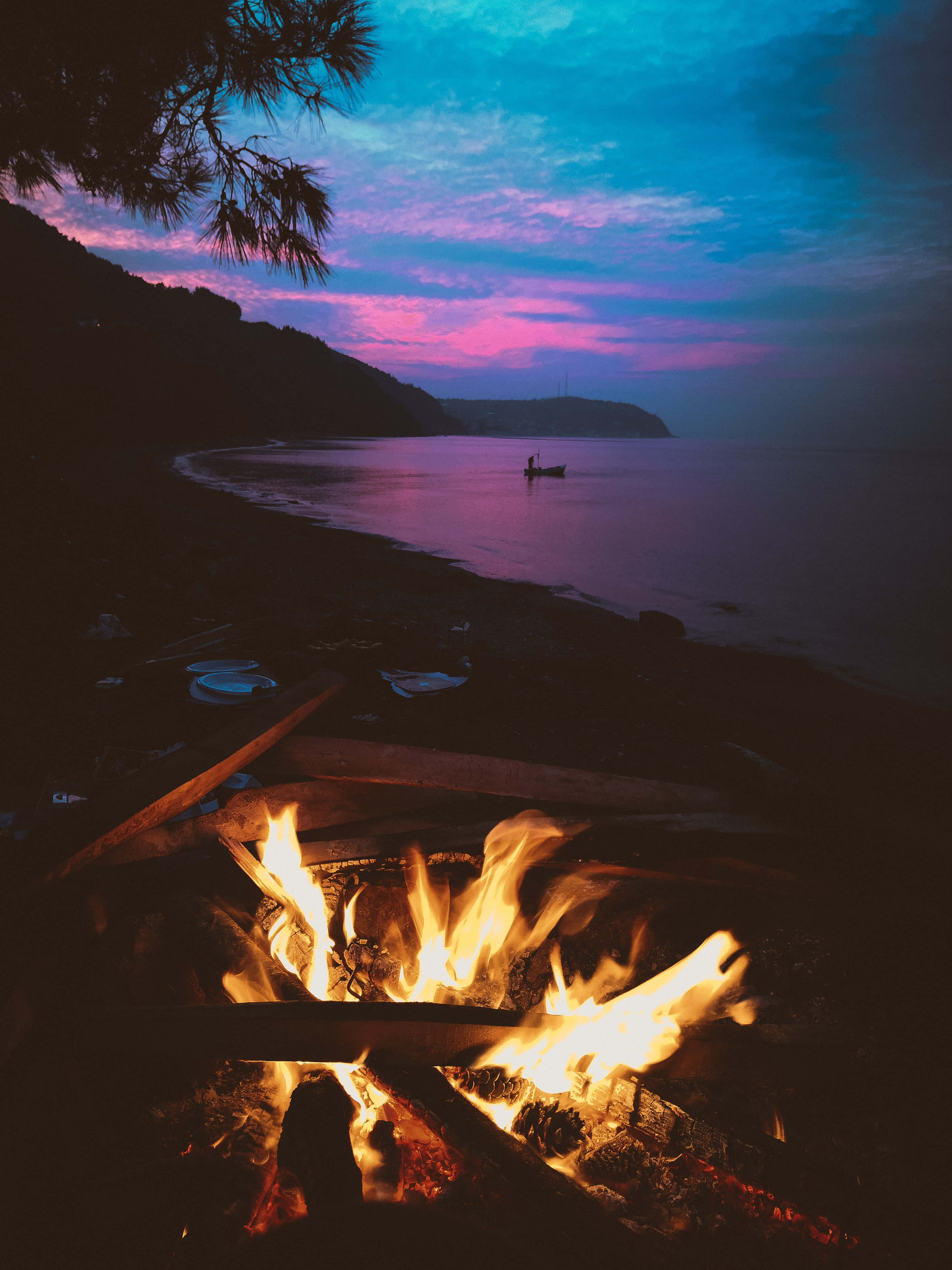 Fall Bonfire 桌布 壁紙 — 一個充滿活力的營火在黃昏的黑暗海灘上噼啪作響，向超現實的粉紅和藍色天空投射溫暖的光芒，一艘孤獨的小船在平靜的水面上漂浮。