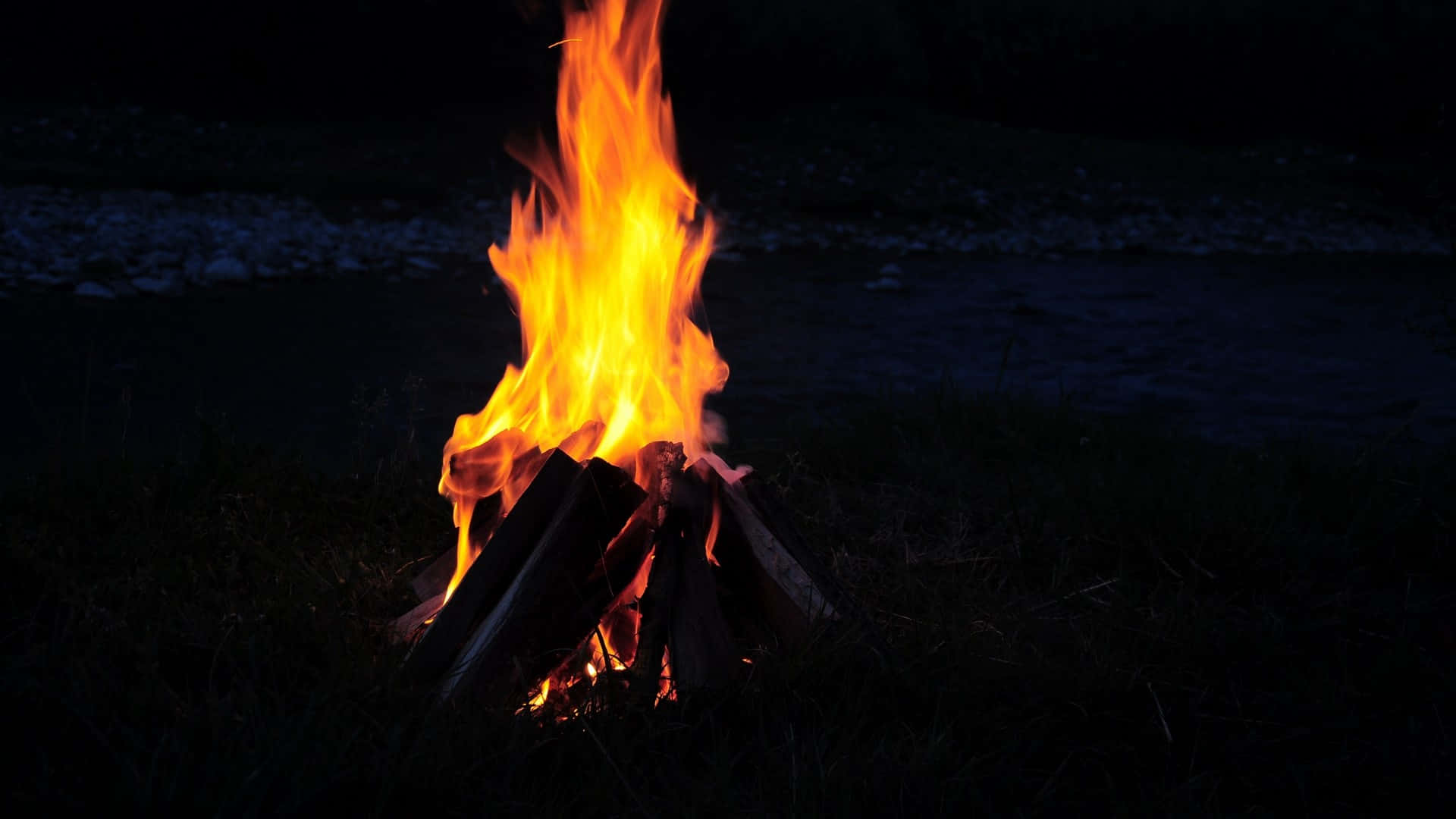 Fall Bonfire 桌布 壁紙 — 一個充滿活力的營火在黑暗的夜晚背景下，以草地和遠處的水面為襯托，發出橙色的火焰噼啪作響。