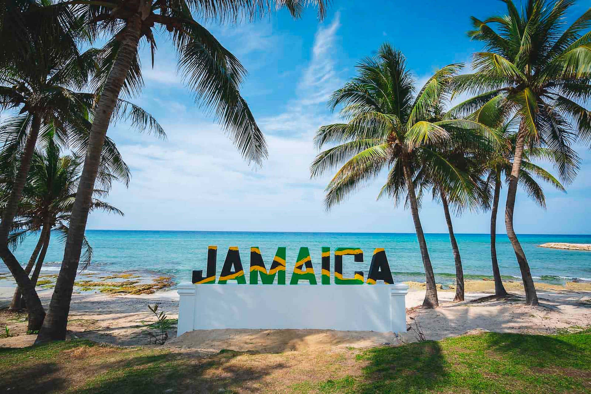 Jamaica 桌布 壁紙 — 一個充滿活力的牙買加標誌矗立在陽光明媚的海灘上，周圍環繞著搖曳的棕櫚樹和碧綠的海洋，天空清澈湛藍。