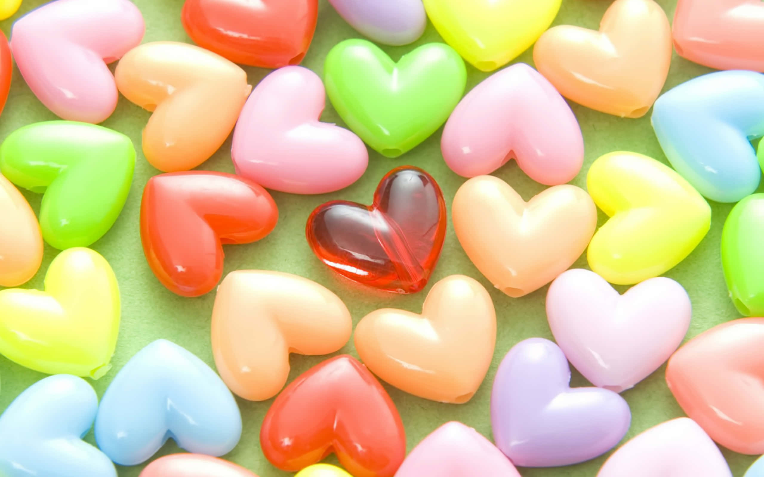 3d Heart 桌布 壁紙 — 一個充滿活力的特寫，呈現出閃亮的心形糖果，以柔和彩虹色調散落在柔軟的綠色表面上。