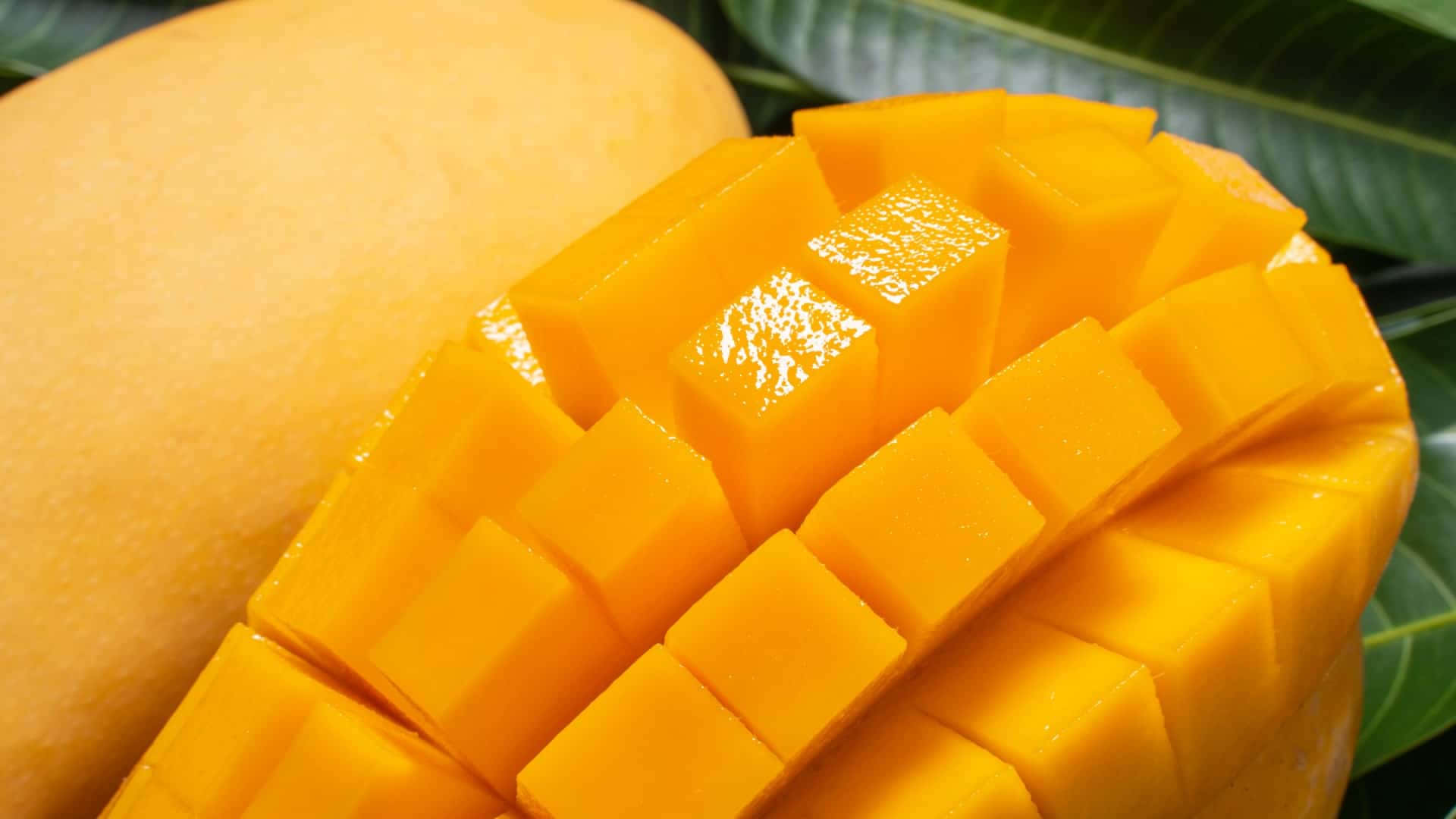 Mango 桌布 壁紙 — 一個充滿活力的特寫，展示成熟的芒果切成多汁的方塊，旁邊是光滑的果皮和茂盛的綠葉。