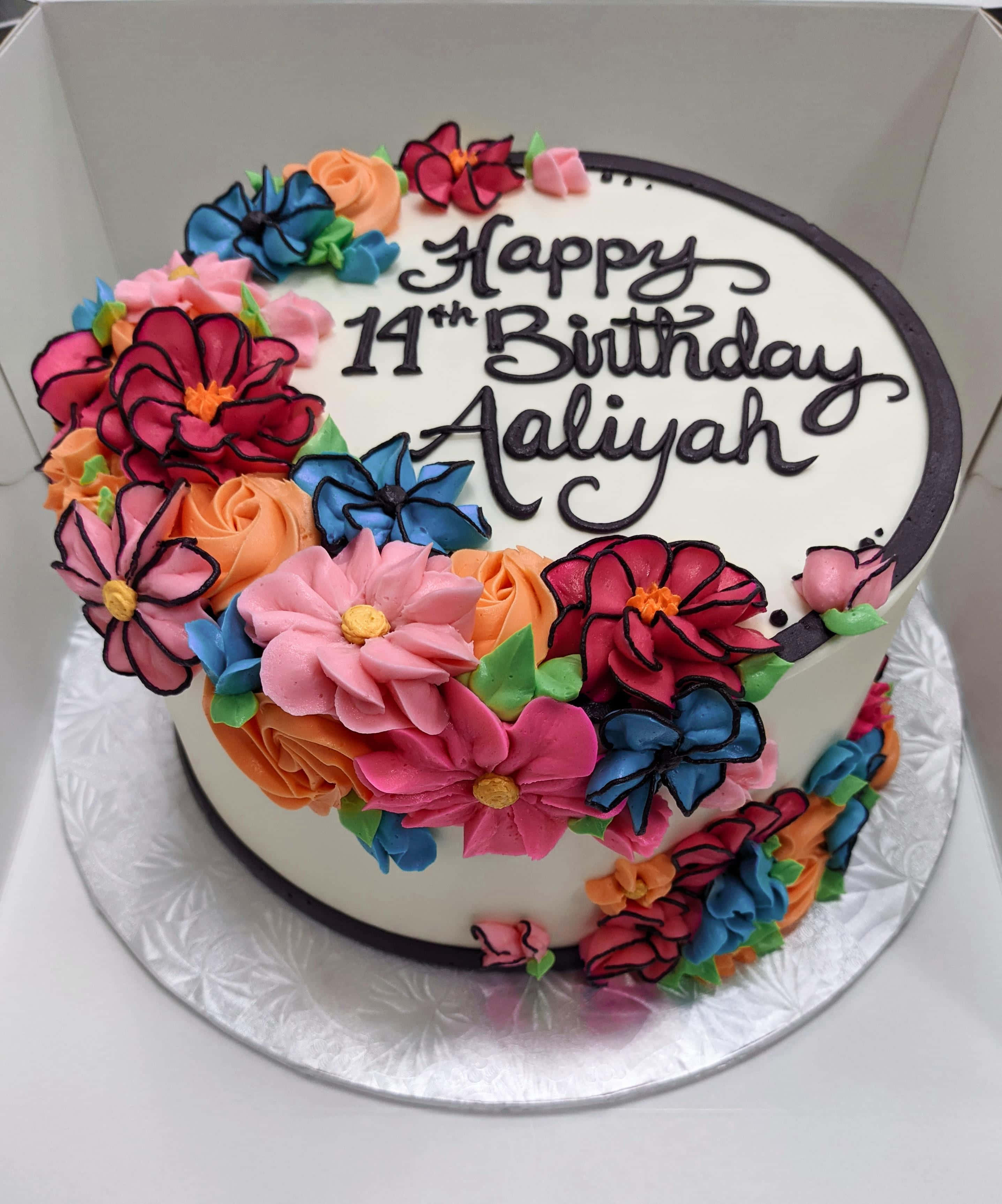 Floral Cake 桌布 壁紙 — 一個充滿活力的生日蛋糕，飾有色彩繽紛的擠花和黑色草書文字「Happy 14th Birthday Aaliyah」，放在一個白色盒子裡。
